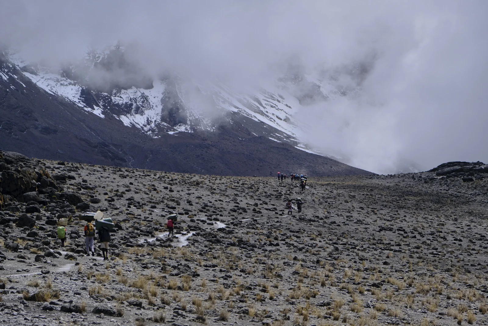 Porters crossing scree toward snowy slopes in cloud