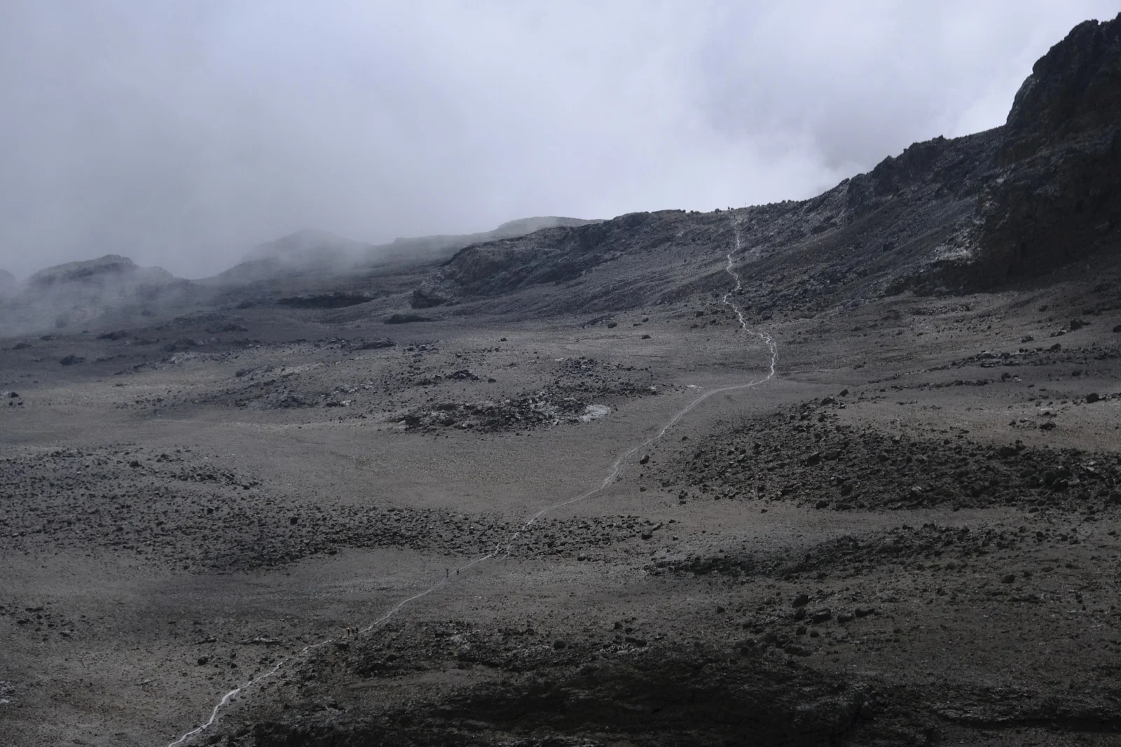 A thin trail crossing a grey alpine slope under cloud