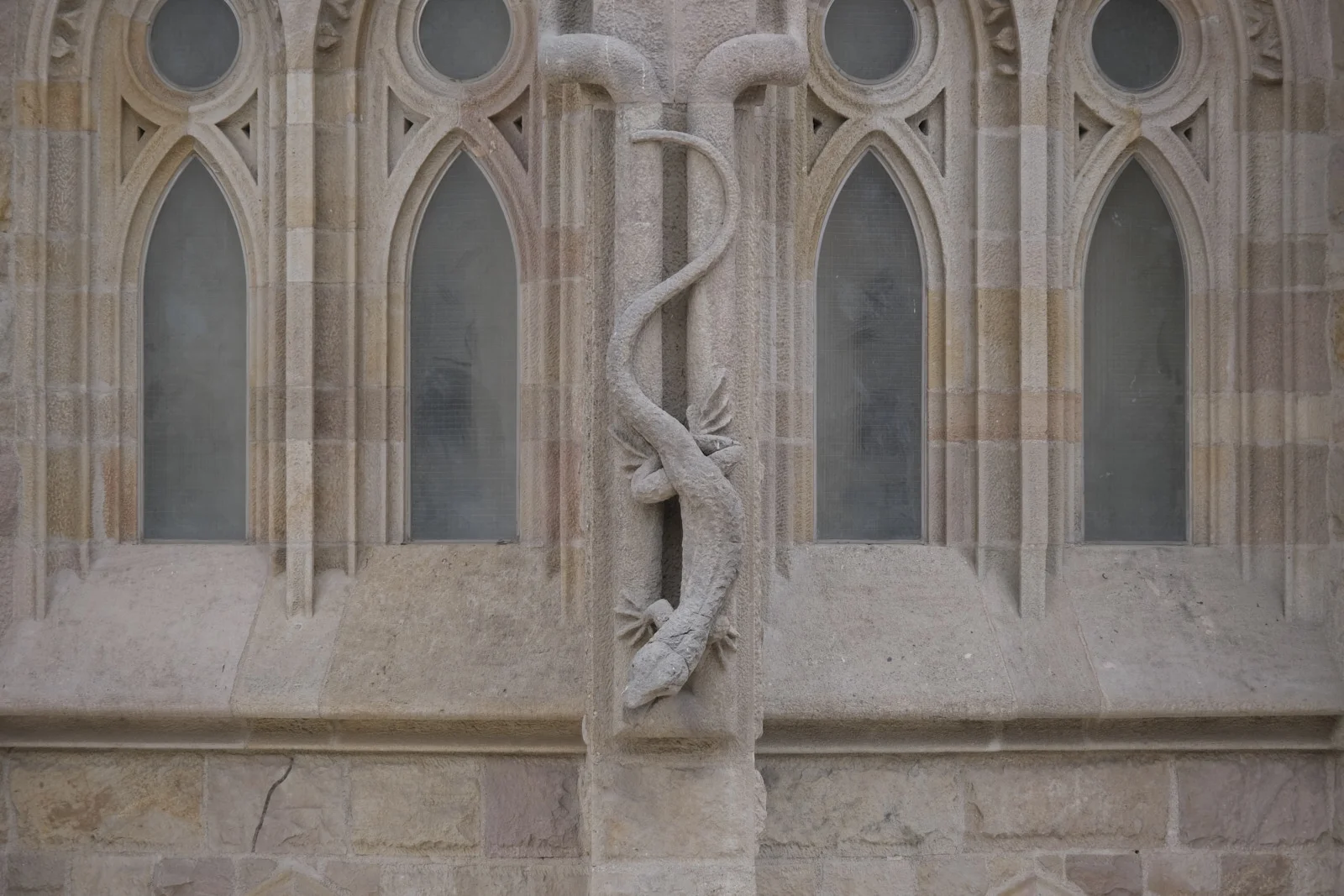 A stone chameleon carved head-down on a column between pointed windows on the Nativity facade of the Sagrada Família in Barcelona.