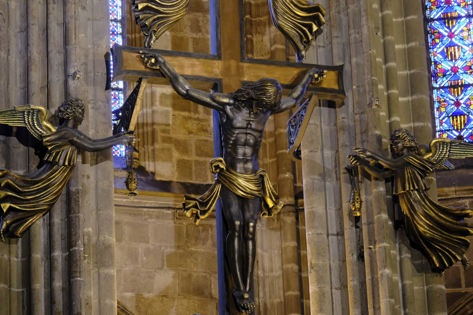 A crucifix flanked by angels holding censers at the high altar of Barcelona Cathedral, with stained glass behind it.