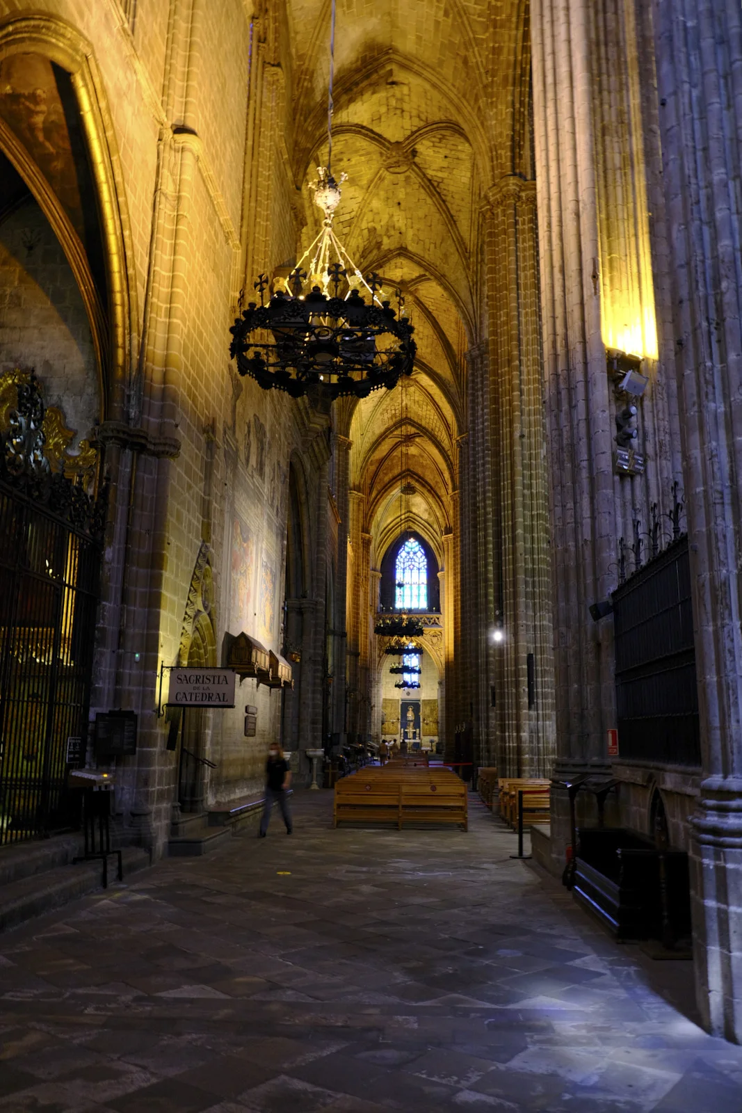 The nave of Barcelona Cathedral, with stone columns, a chandelier, pews, and a sign reading Sacristia de la Catedral.