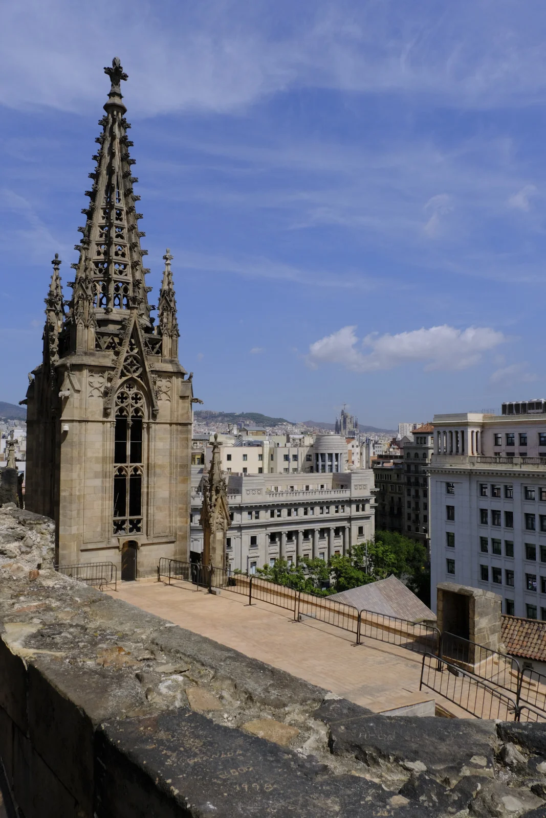 A gothic stone spire on the roof terrace of Barcelona Cathedral, with the Sagrada Família visible past the surrounding buildings.