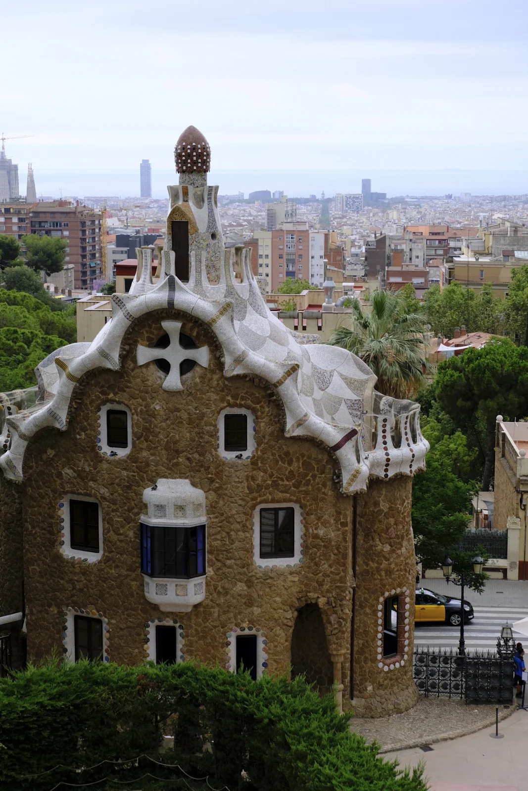 The smaller entrance pavilion at Park Güell in Barcelona, a stone building with a mosaic roof and a cross, above a street with the city beyond.