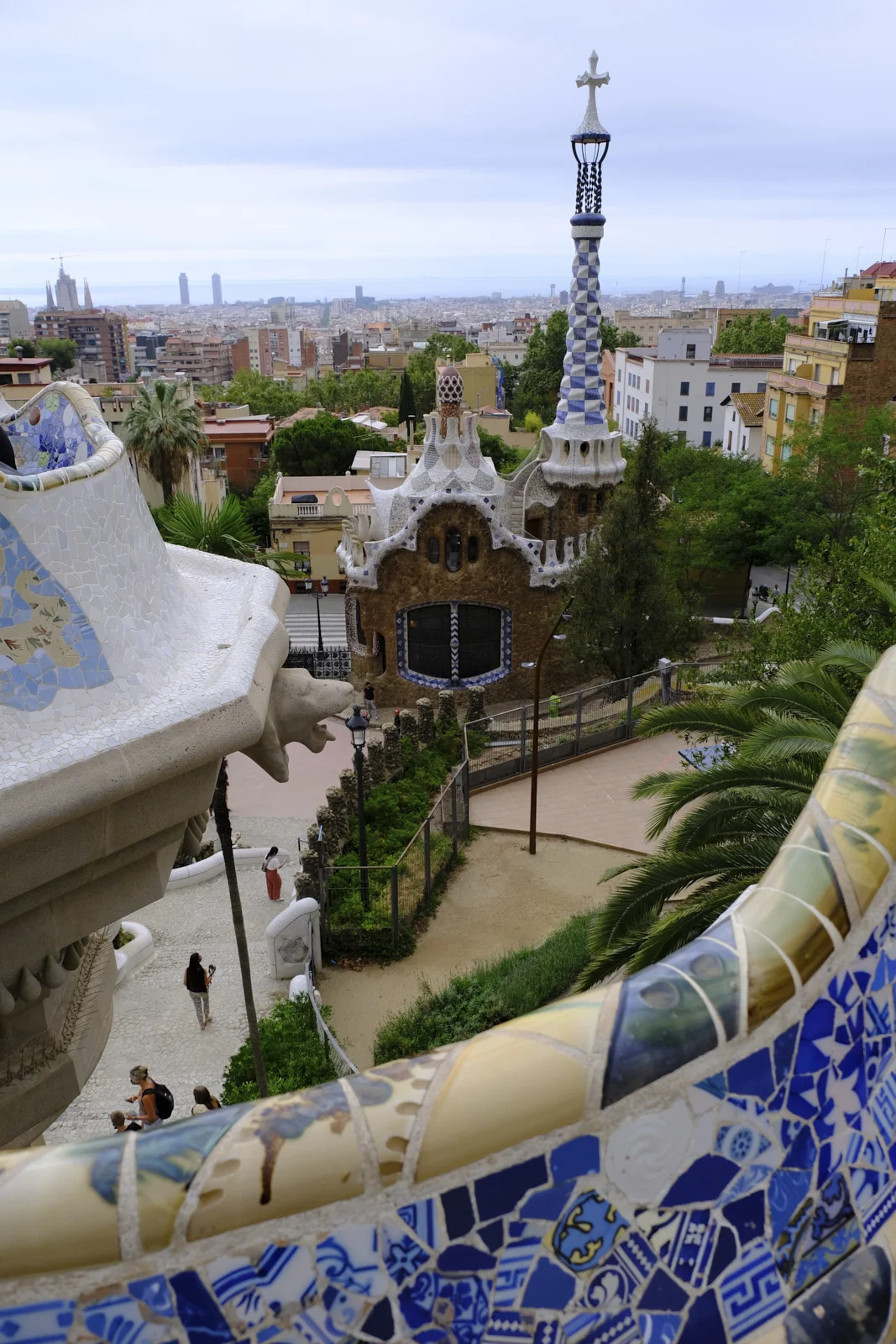 The mosaic bench at Park Güell in Barcelona in the foreground, with a carved stone head and the entrance pavilion's tall spire beyond.