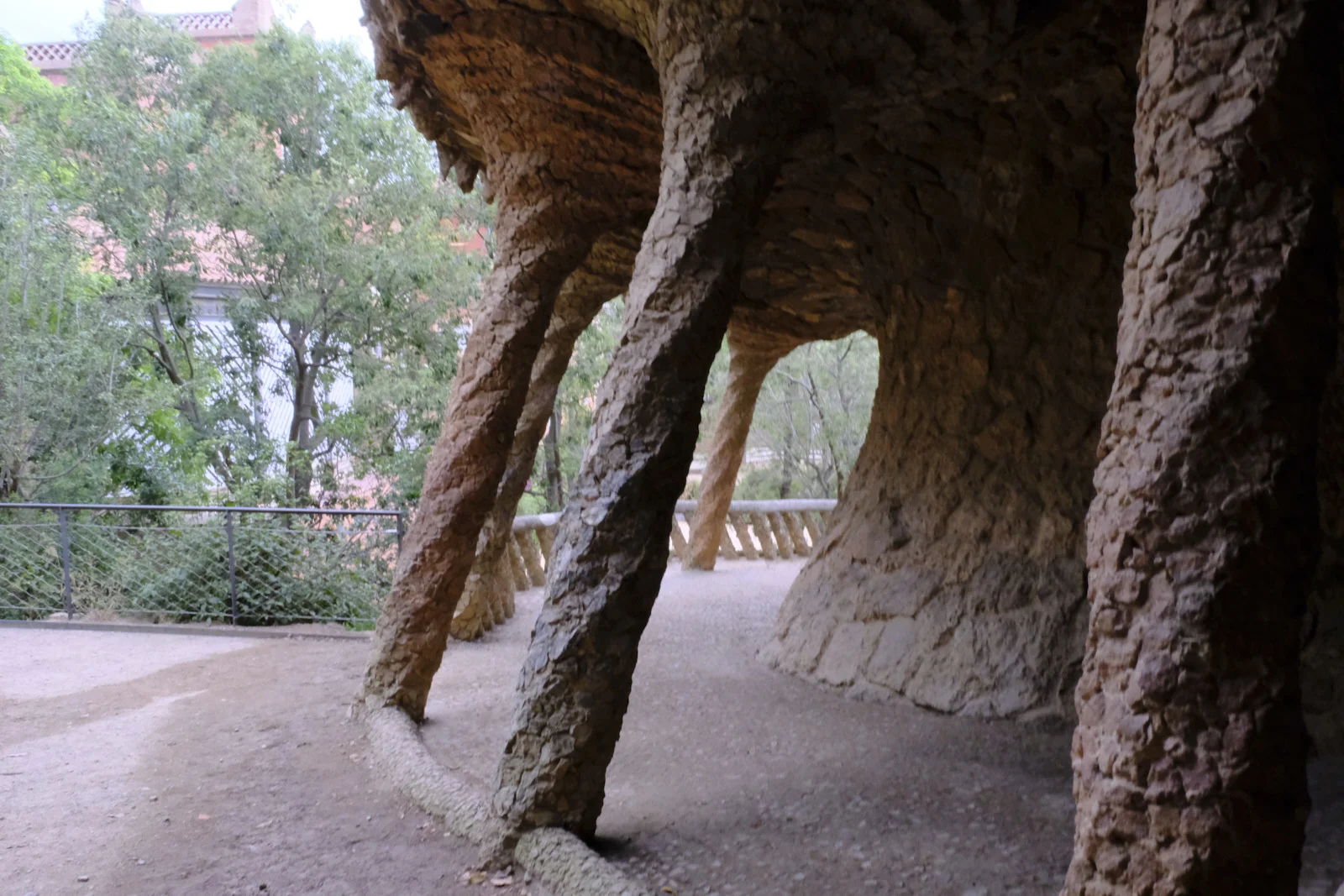 Slanted rough-stone columns under a rock-like vault of a viaduct at Park Güell in Barcelona, opening toward trees.
