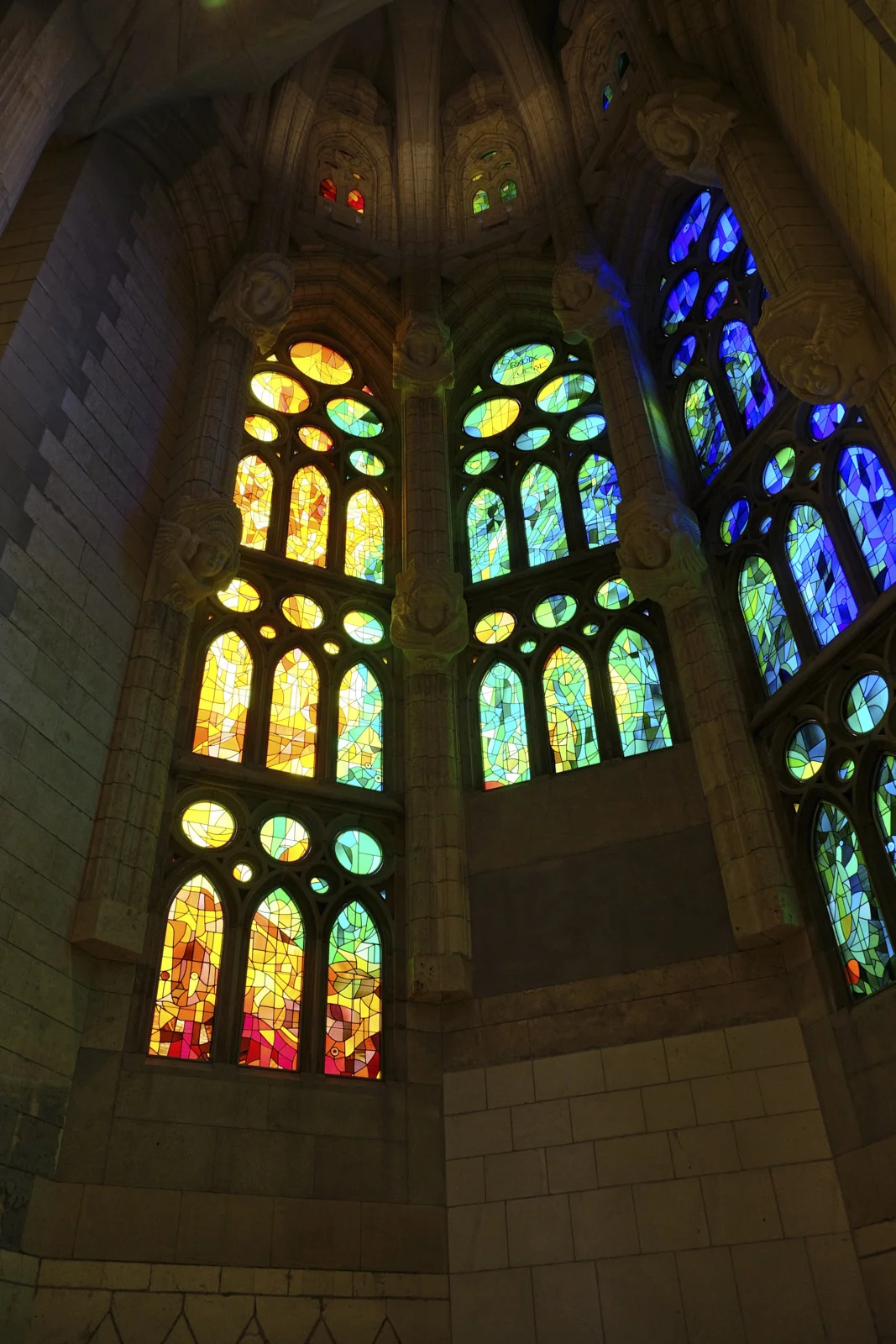 Tall stained-glass windows divided by stone columns with carved heads in the nave of the Sagrada Família in Barcelona.