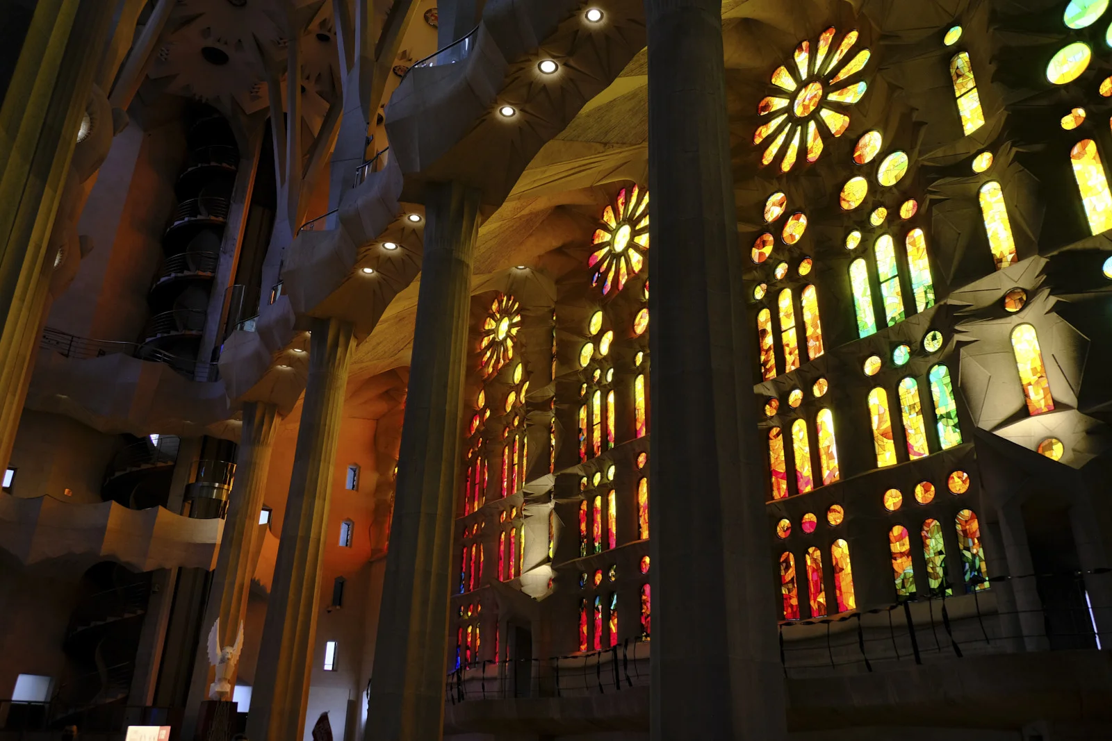 Branching stone columns and tall stained-glass windows inside the Sagrada Família in Barcelona, with a spiral stair in the tower at left.