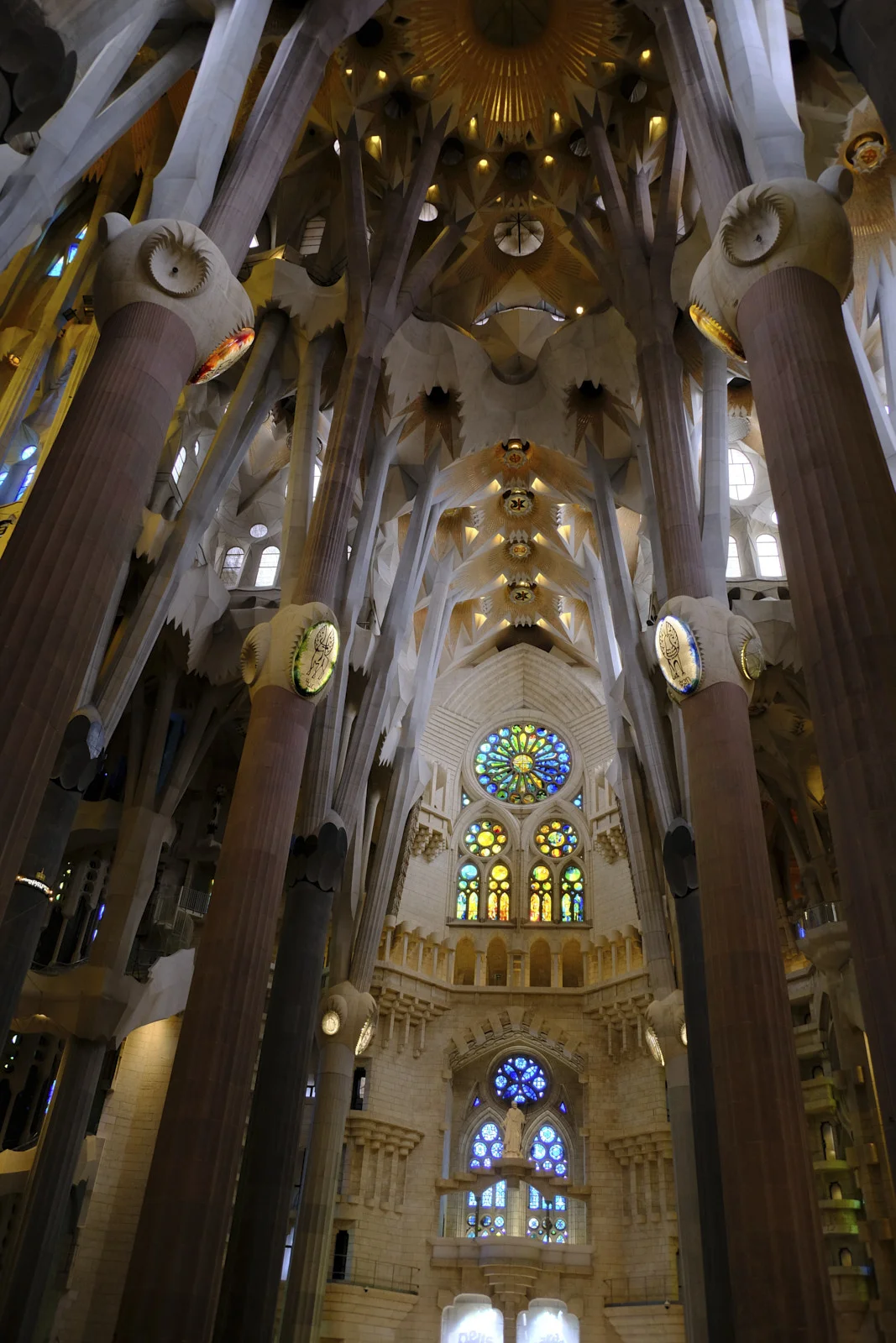 Branching columns and vaults of the Sagrada Família in Barcelona, looking toward stacked rose windows and a statue on the Nativity facade.
