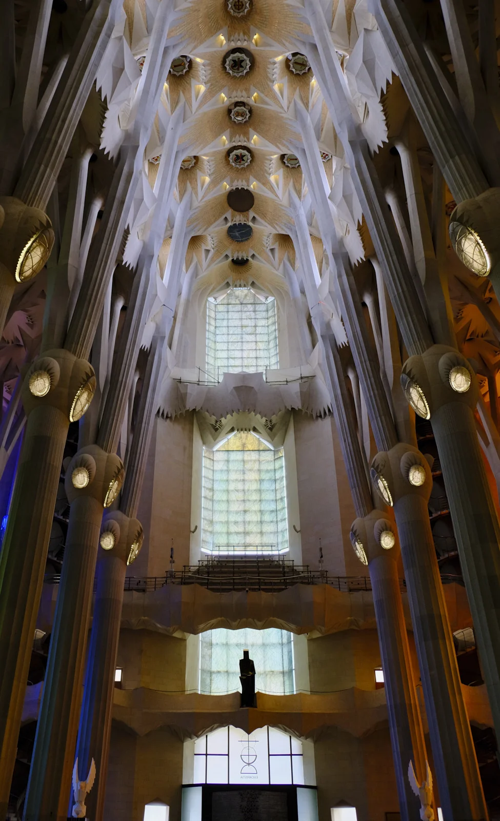 The nave columns and vaults of the Sagrada Família in Barcelona, with a dark standing figure in front of tall plain windows.