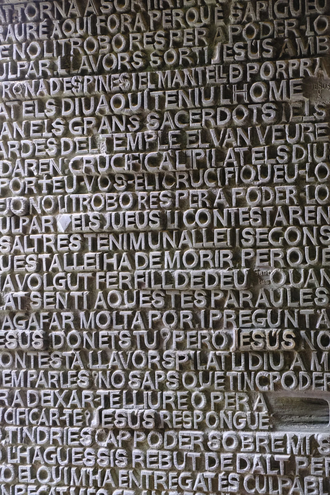 A close view of raised Catalan letters covering a bronze door on the Passion facade of the Sagrada Família in Barcelona, including the names Jesús and Pilat.