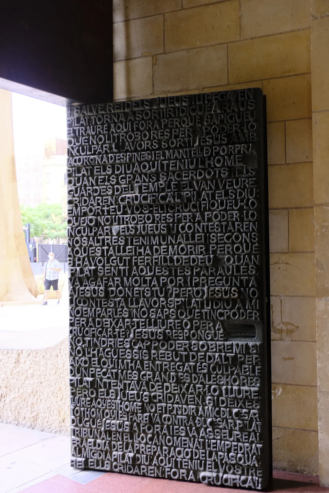 A tall bronze door covered in raised Catalan text at the Passion facade of the Sagrada Família in Barcelona, set in a stone doorway.
