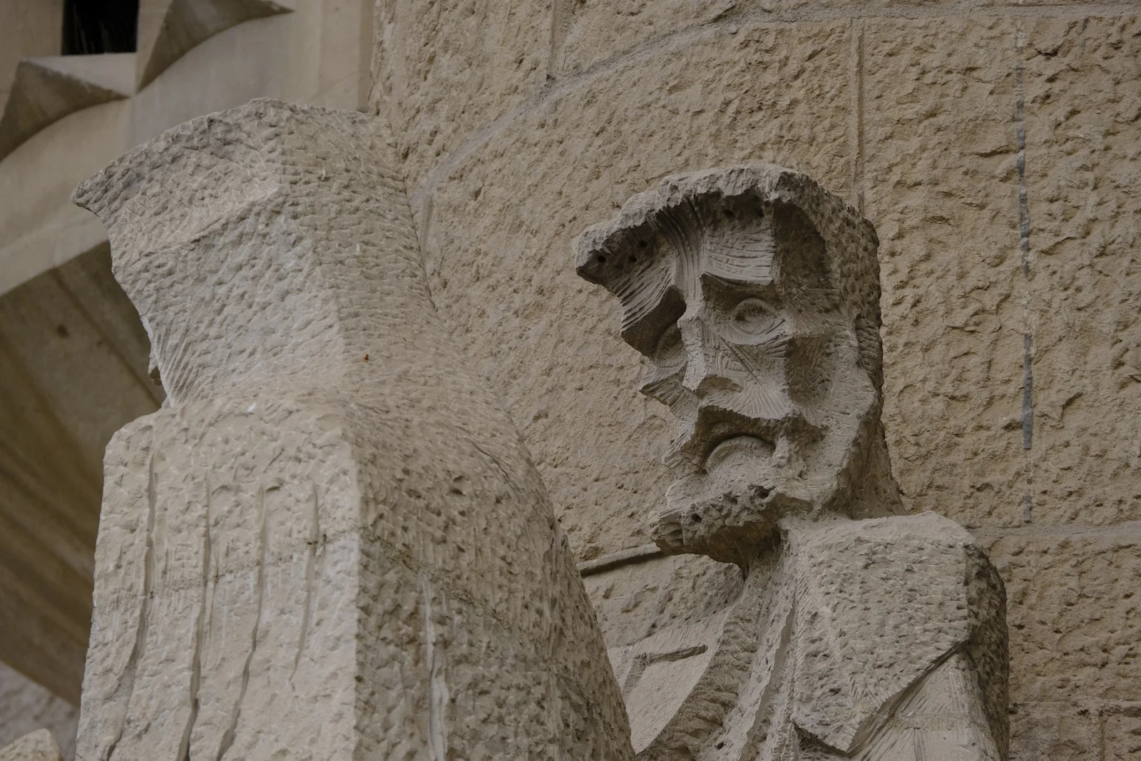 A bearded stone head on the Passion facade of the Sagrada Família in Barcelona, beside a hooded figure.