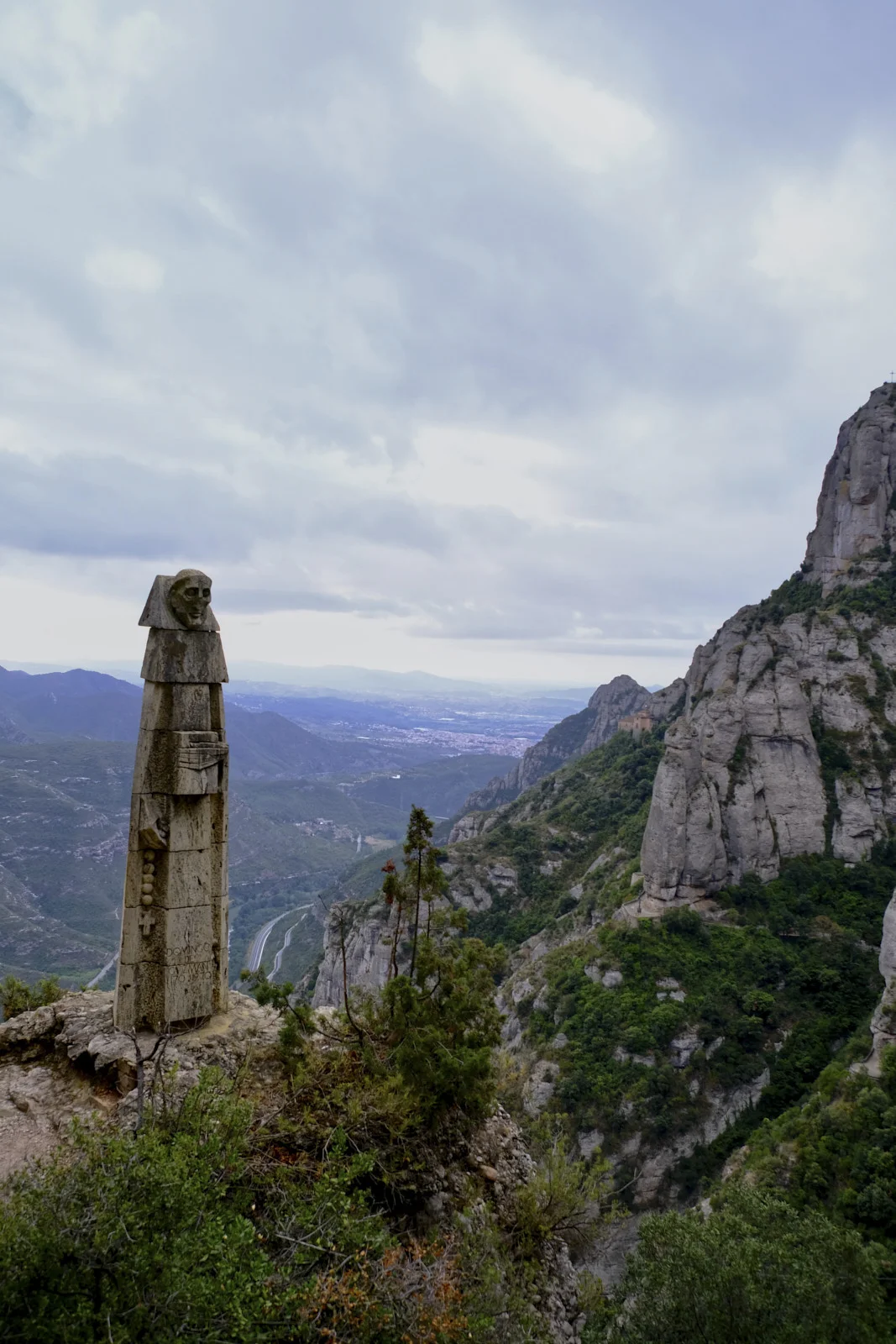 A tall stacked-stone statue of Sant Domènec, face and hands emerging from the block and a rosary on the side, on the path to Santa Cova at Montserrat.