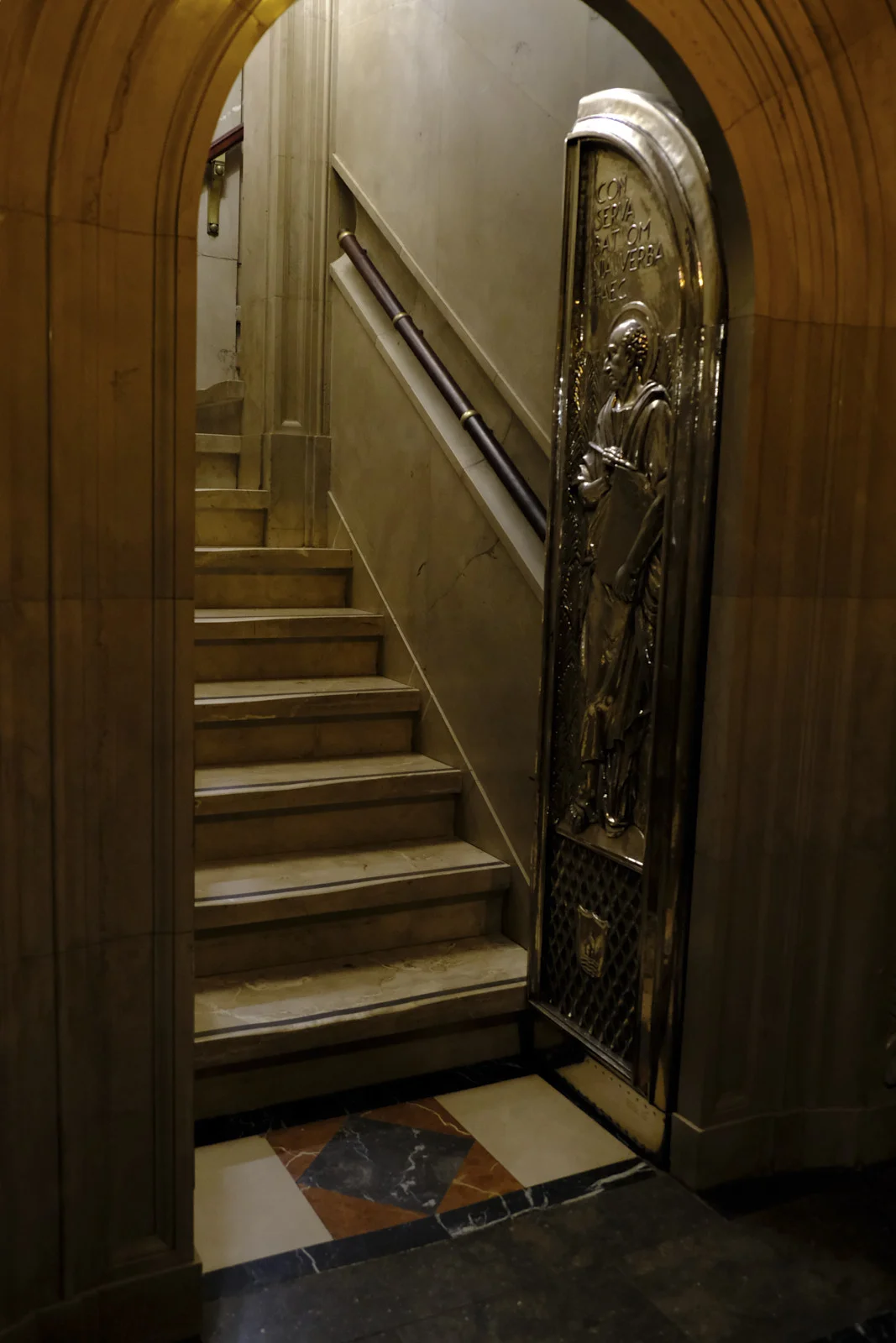 A bronze door with a relief of a haloed saint beside a marble stair in the basilica at Montserrat.