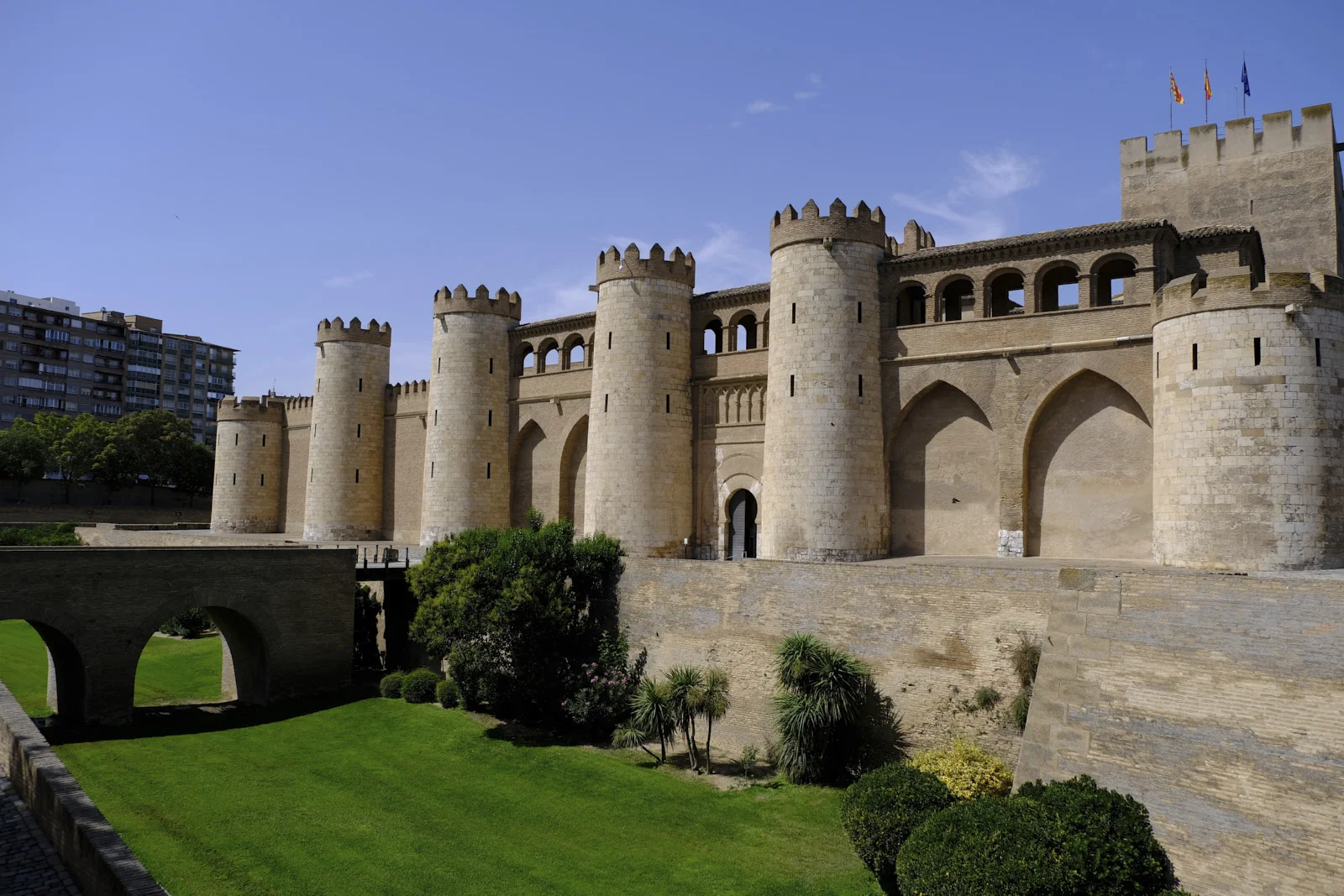 Round towers and the square keep of the Aljafería Palace in Zaragoza rise above a moat, with an arched bridge at the left.