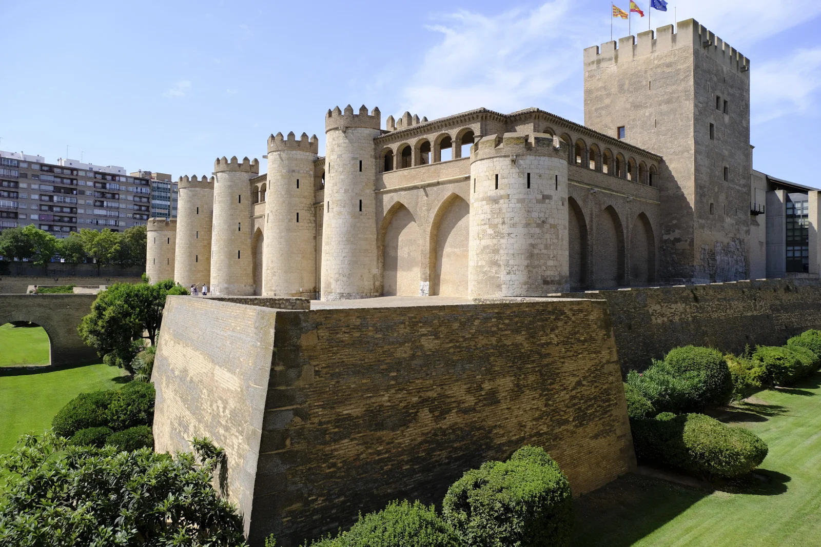 A row of round towers and the square keep of the Aljafería Palace in Zaragoza stand above a brick rampart.