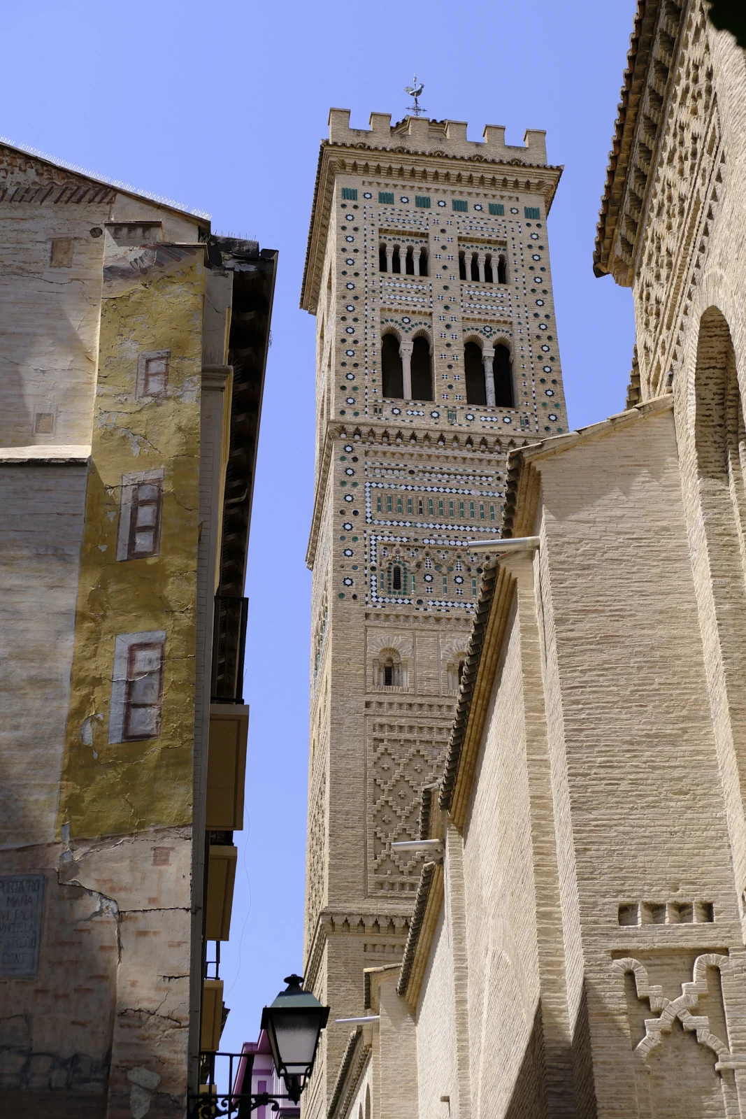 The square bell tower of the Church of La Magdalena in Zaragoza, topped by a rooster weathervane, rises between the surrounding buildings.