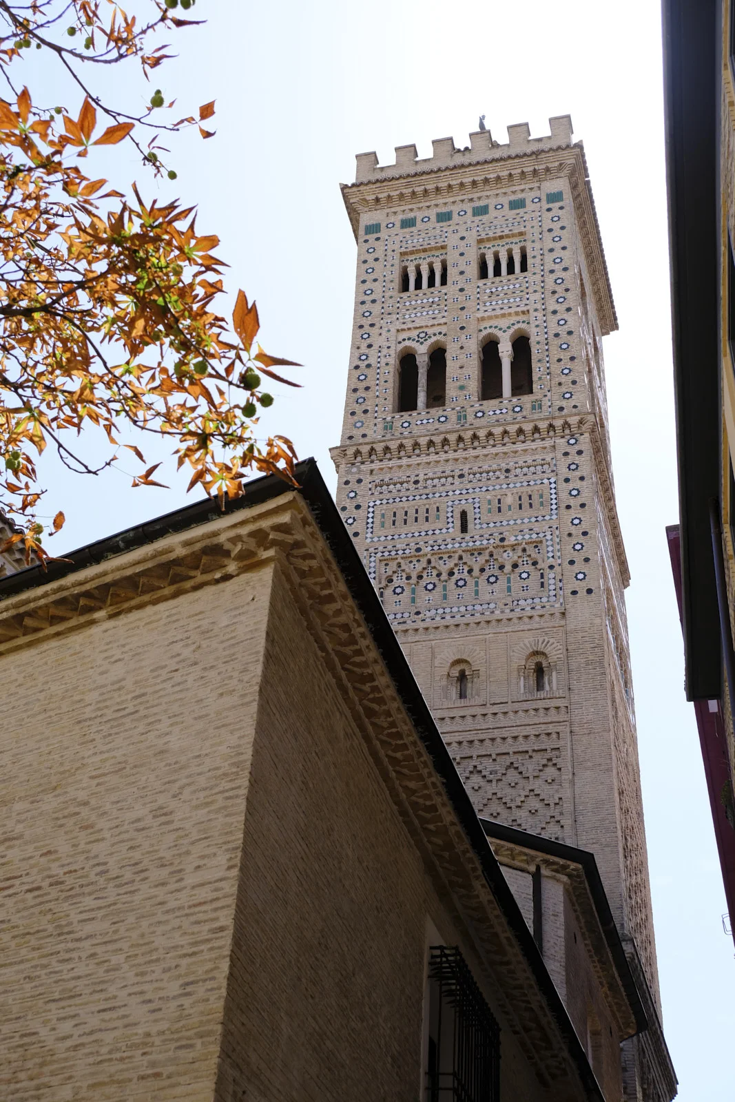 The Mudéjar bell tower of the Church of La Magdalena in Zaragoza is seen from street level above a nearby roof.