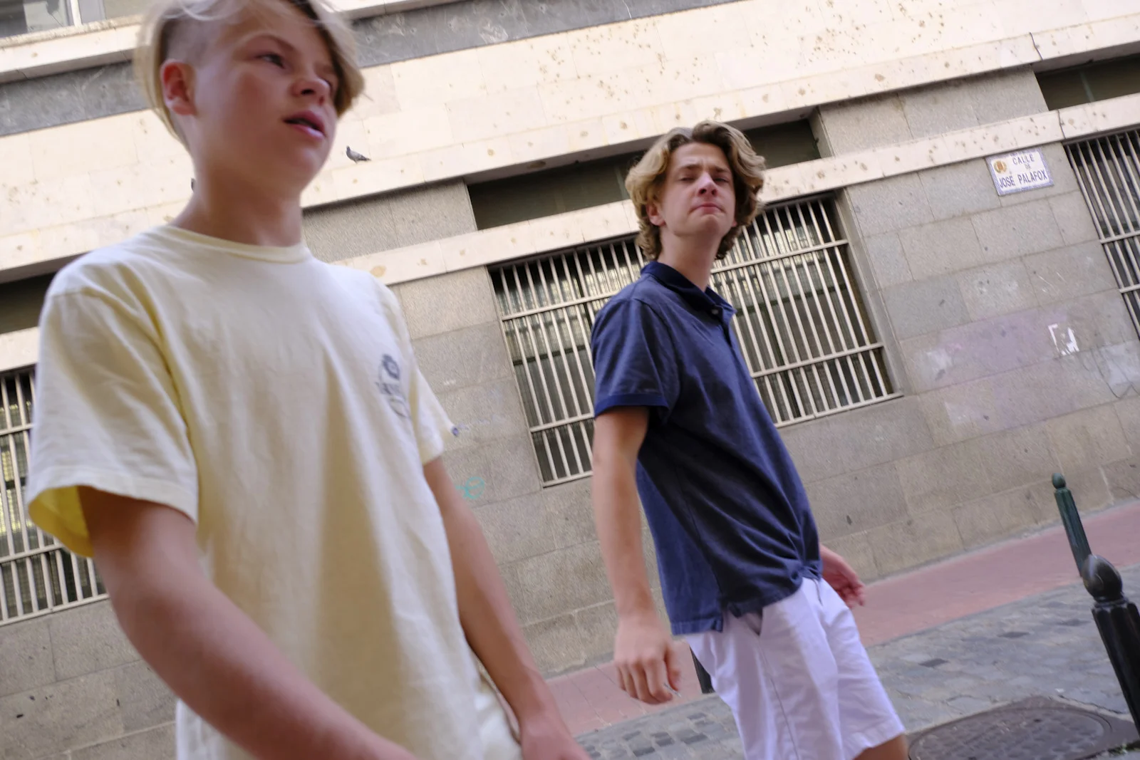Two boys stand on Calle José Palafox in Zaragoza, under a street sign on a stone building with barred windows.