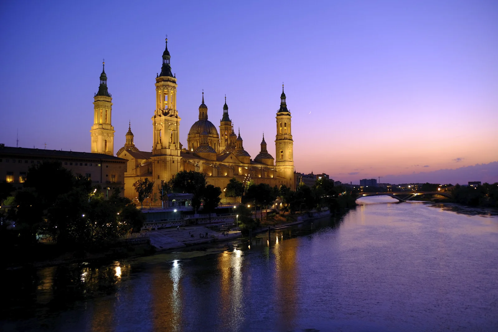 The towers and domes of the Basilica of Our Lady of the Pillar in Zaragoza rise beside the Ebro, with a bridge farther along the river.