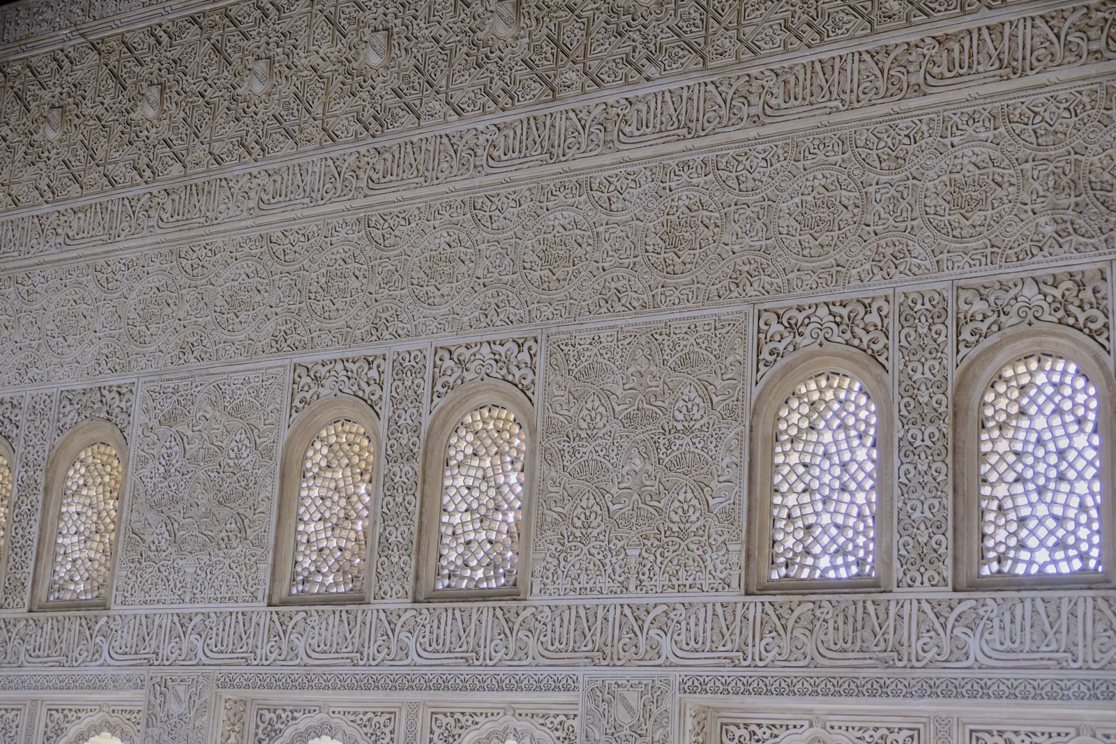 A long wall of carved plaster and Arabic inscription bands runs above and below a row of arched lattice windows.