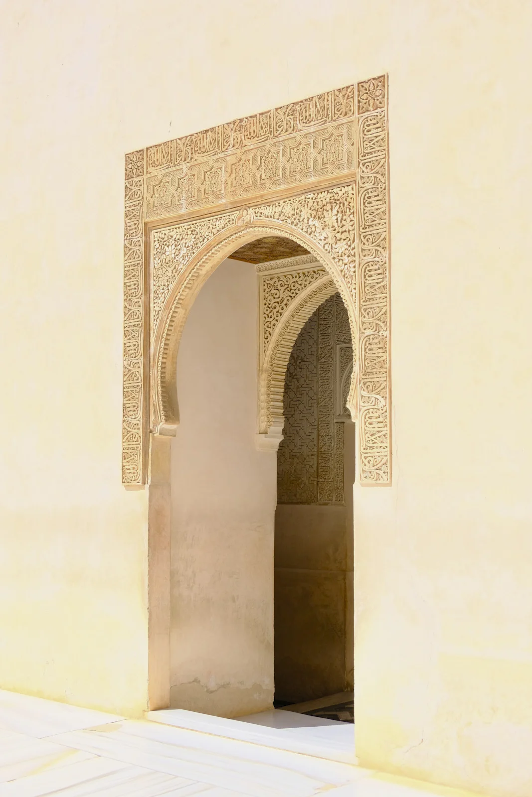 A horseshoe arch carved with geometric panels and Arabic script opens through a plain wall onto another carved arch.