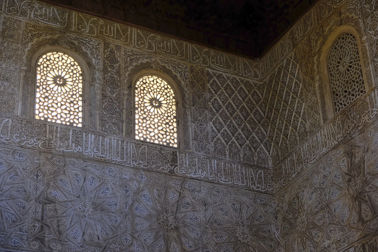 Three arched lattice windows sit in an upper wall of Arabic inscriptions, star patterns, and geometric plaster carving.