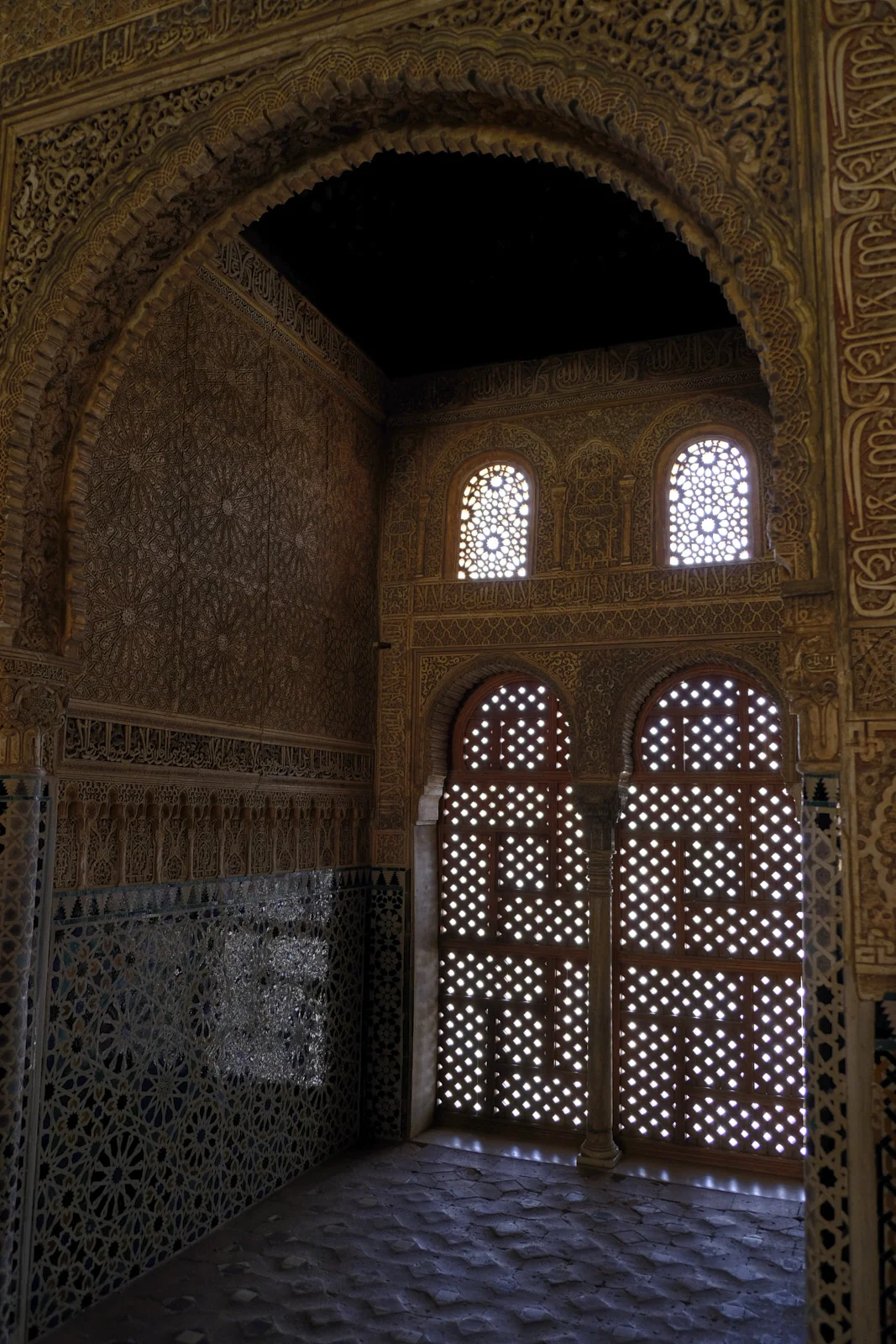 An arch frames a chamber whose far wall has two tall wooden lattice screens, smaller lattice windows above them, carved stucco, and a geometric tile dado.