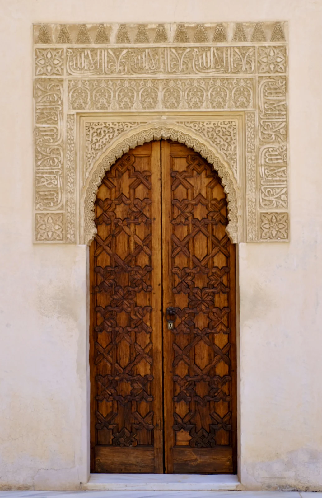 Double wooden doors carved with an interlaced geometric pattern stand under a stucco horseshoe arch and a panel of Arabic inscription.