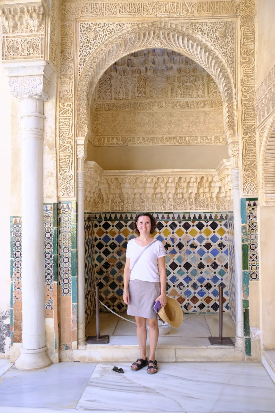 A woman stands in an alcove lined with geometric tile under a carved stucco horseshoe arch.