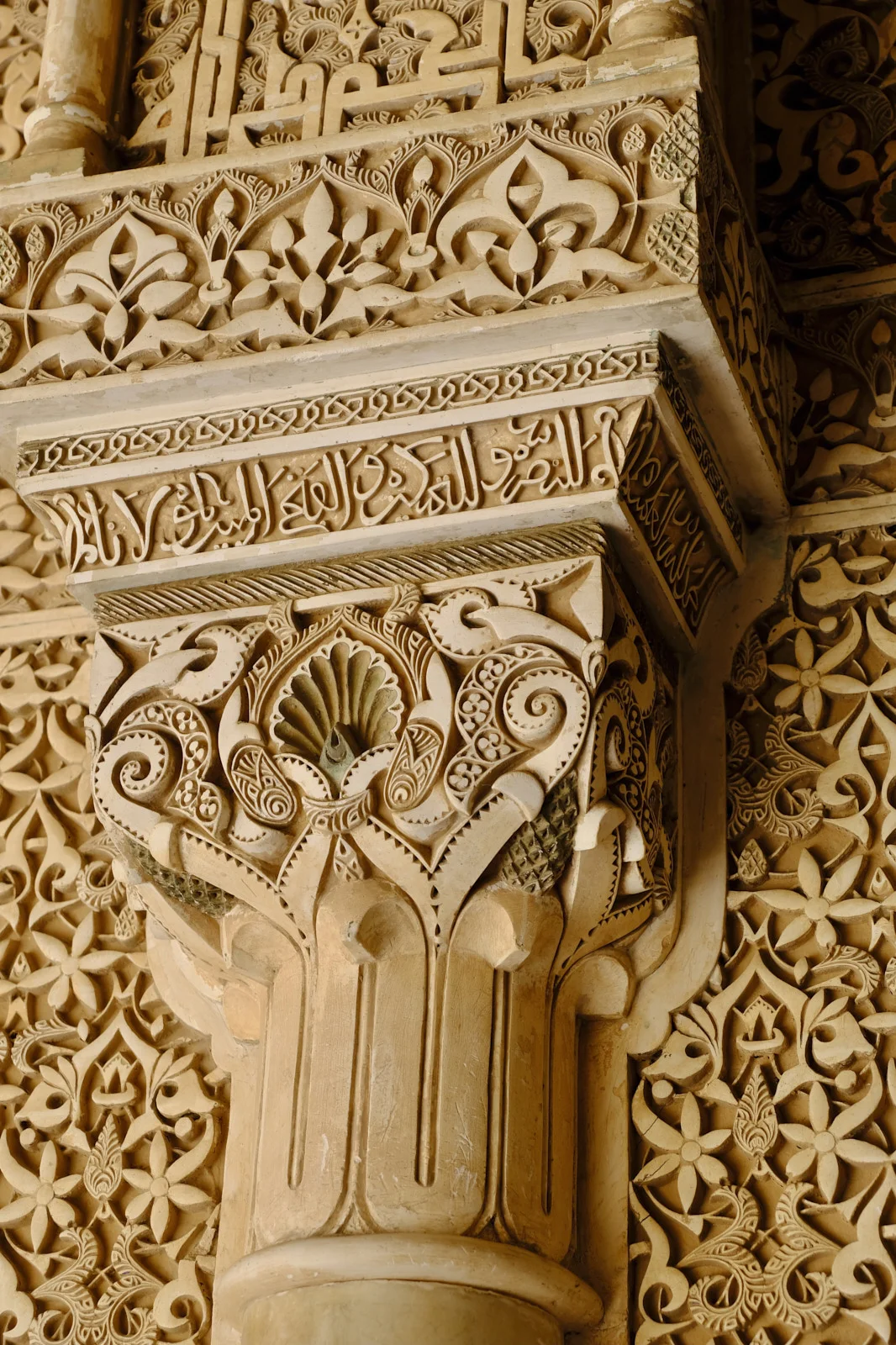 A column shaft and a carved capital with a shell, scrolls, and Arabic script on the abacus, against a wall of stucco.
