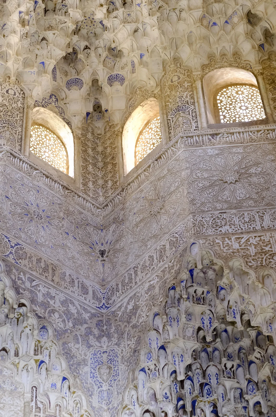 Lattice windows, a geometric star panel, and tiers of muqarnas meet in a corner of a palace hall.