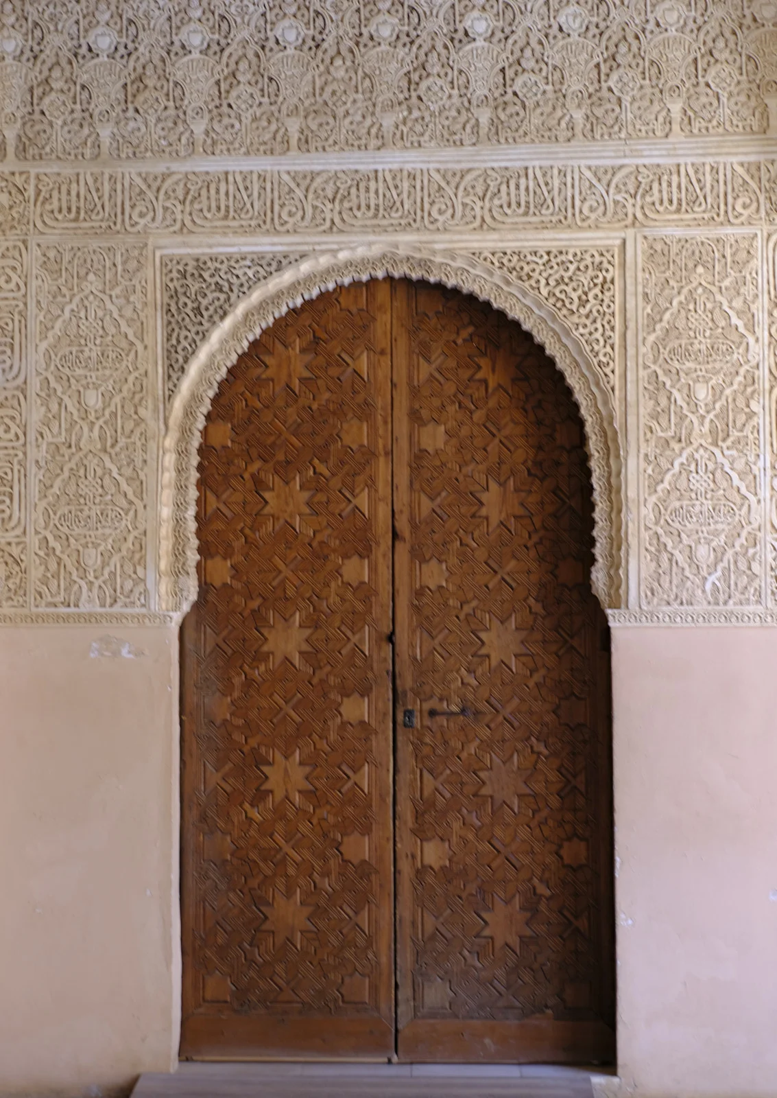 Double wooden doors covered with eight-pointed stars sit under a stucco arch and bands of carved inscription.