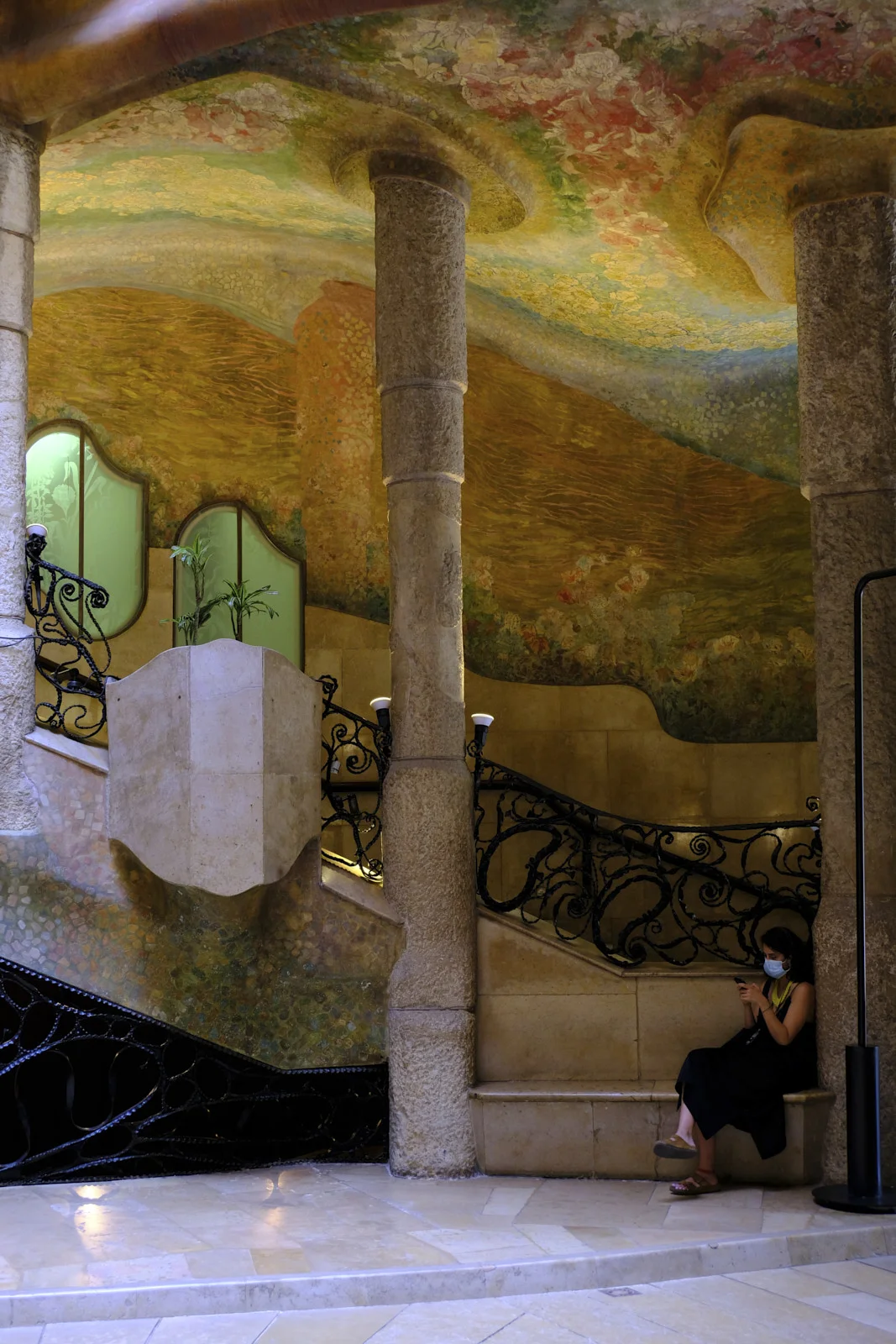 Stone columns, an iron stair, and a painted ceiling fill the entrance hall of La Pedrera in Barcelona, where a person sits on a bench.