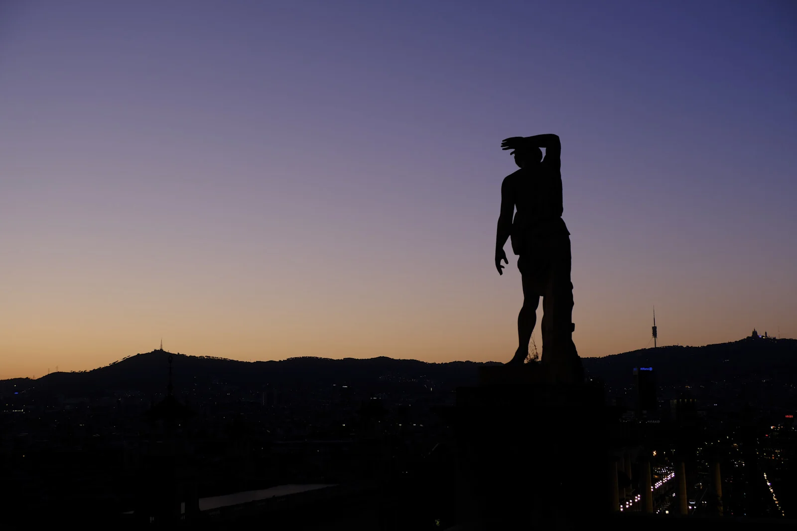 A statue of a standing figure, one hand raised to the forehead, overlooks Barcelona from a terrace.