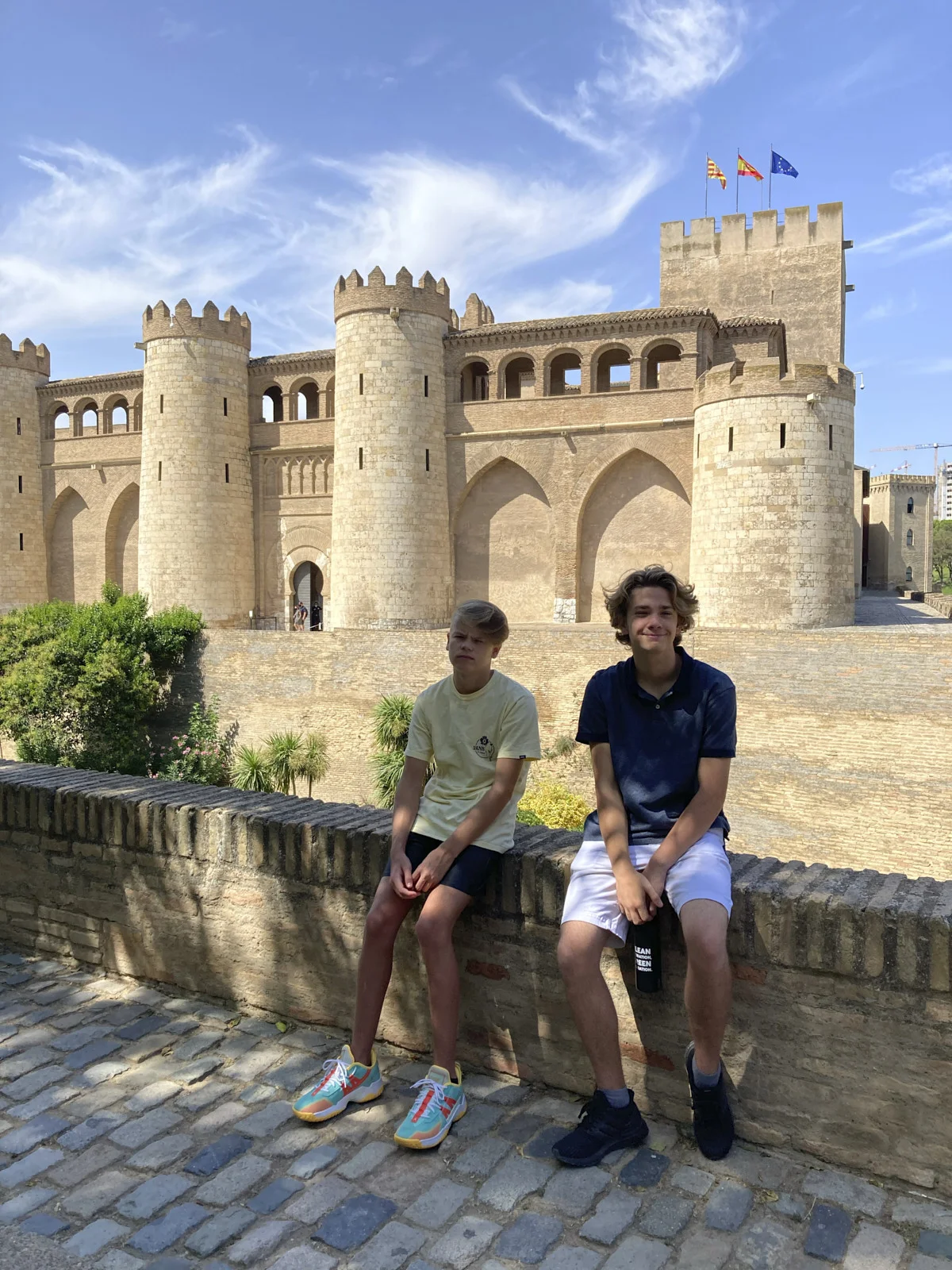 Two boys sit on a brick wall before the round towers and keep of the Aljafería Palace in Zaragoza.