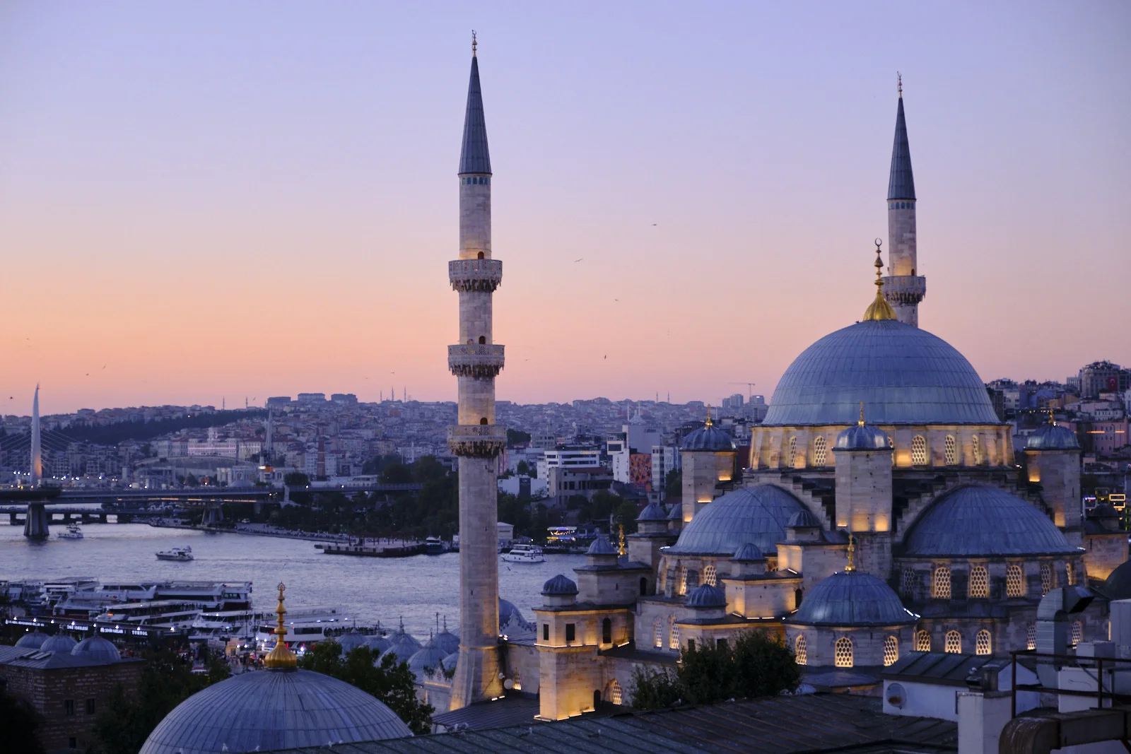 The New Mosque, its domes and minarets lit at twilight, rises beside the Golden Horn with Galata Bridge and ferries beyond.