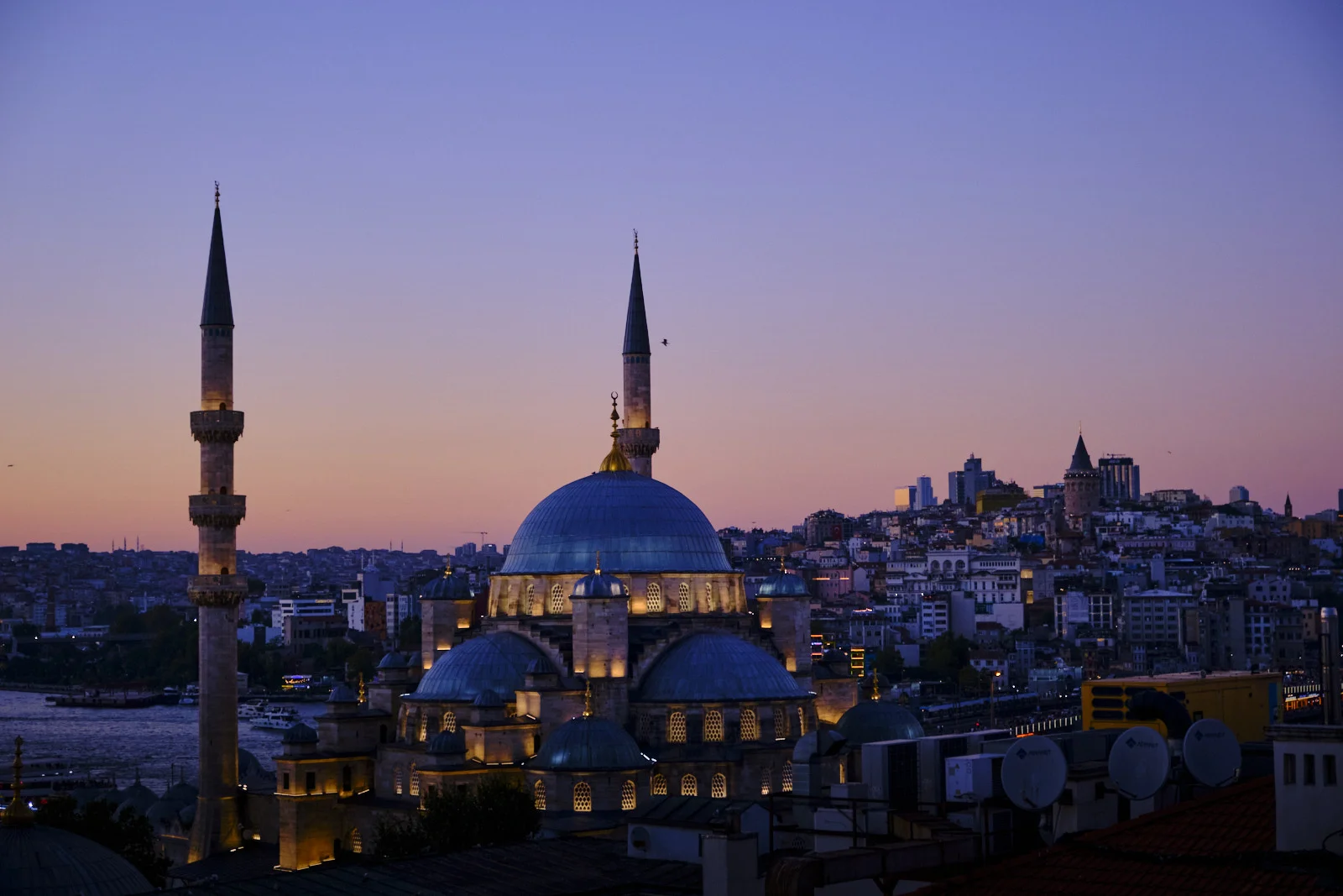The Süleymaniye Mosque at dusk, its dome and minarets lit, with the Golden Horn at lower left and the Galata Tower on the distant hill.