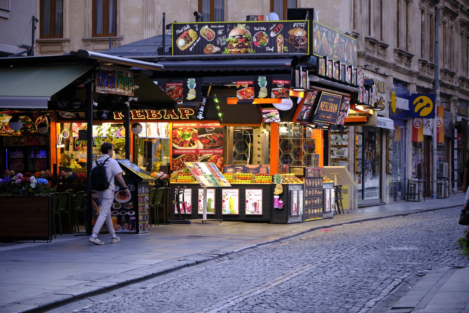 A lit kebab and fresh-juice stall on a cobblestone street, with fruit on display and a man wearing a backpack walking past.
