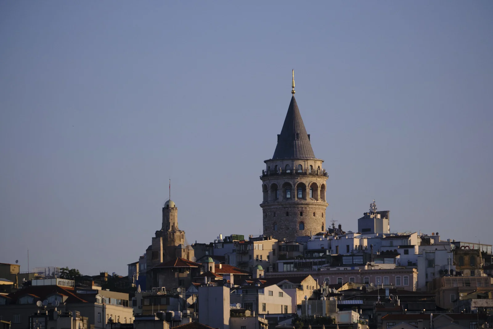 The Galata Tower above dense rooftops in morning light, with a smaller domed tower and a flag to its left.