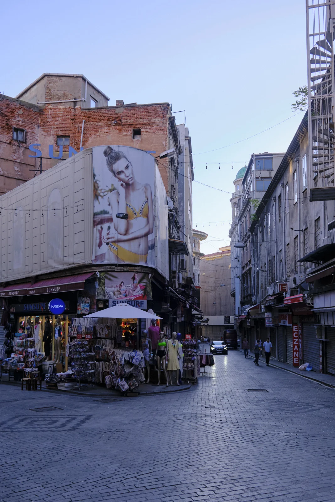 A cobblestone street corner with a large swimsuit billboard, shop mannequins, and closed metal shutters.