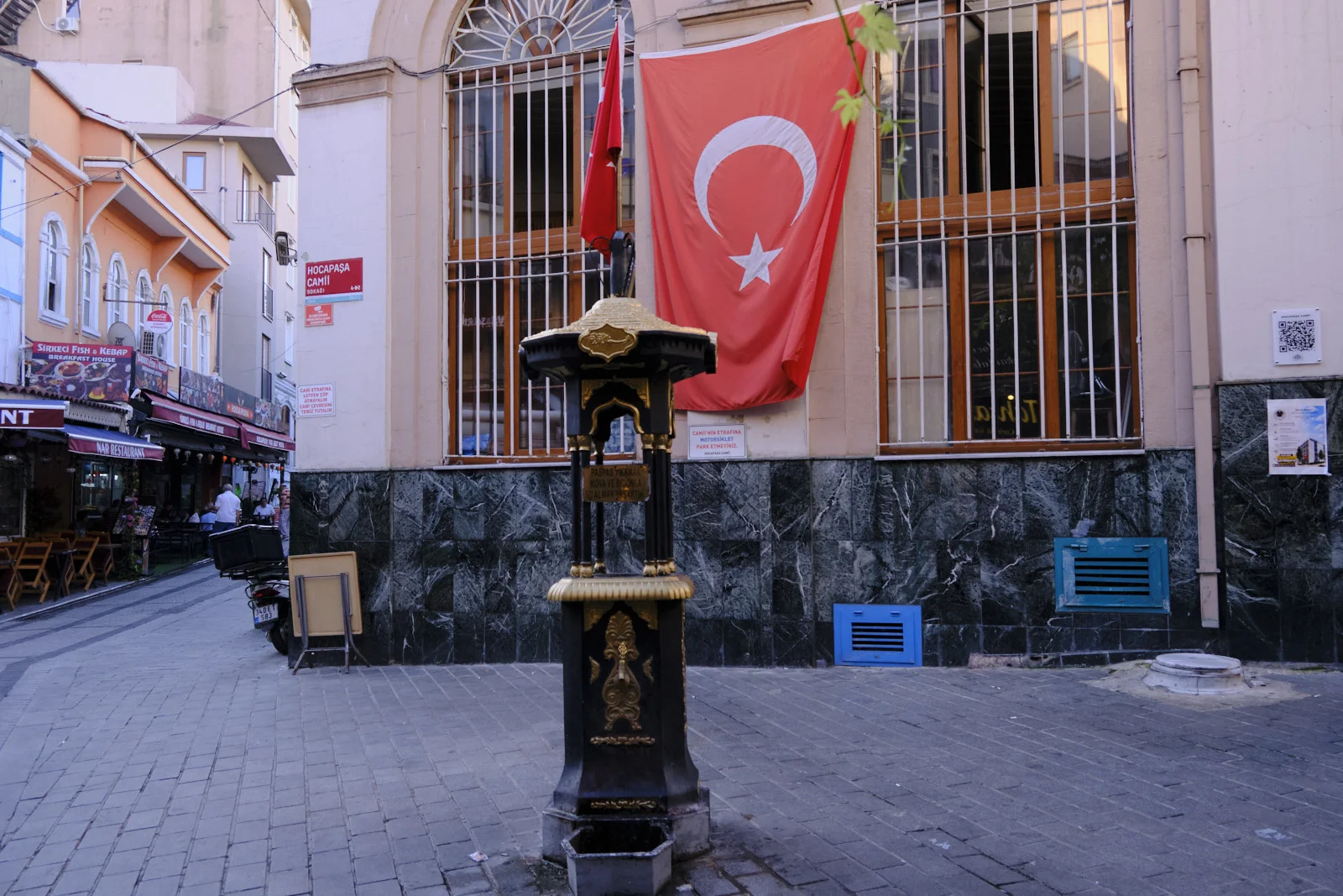 A black and gold Ottoman fountain stands on a paved Sirkeci street beneath a large Turkish flag.