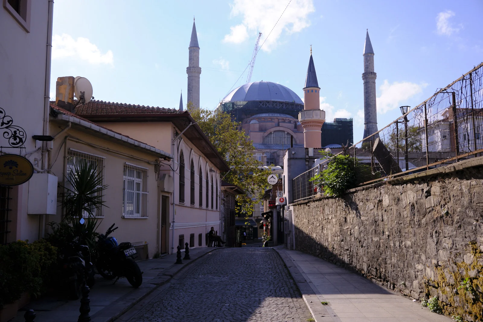 A cobblestone lane leading to Hagia Sophia, its dome wrapped in scaffolding with a crane behind it and motorcycles parked beside the houses.