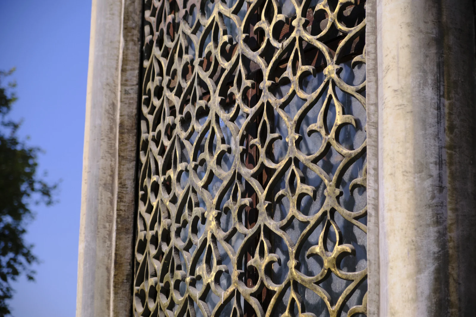 Close view of an ornate gilded window grille with a repeating leaf-and-flower pattern, set in a weathered stone frame with blue sky beyond.