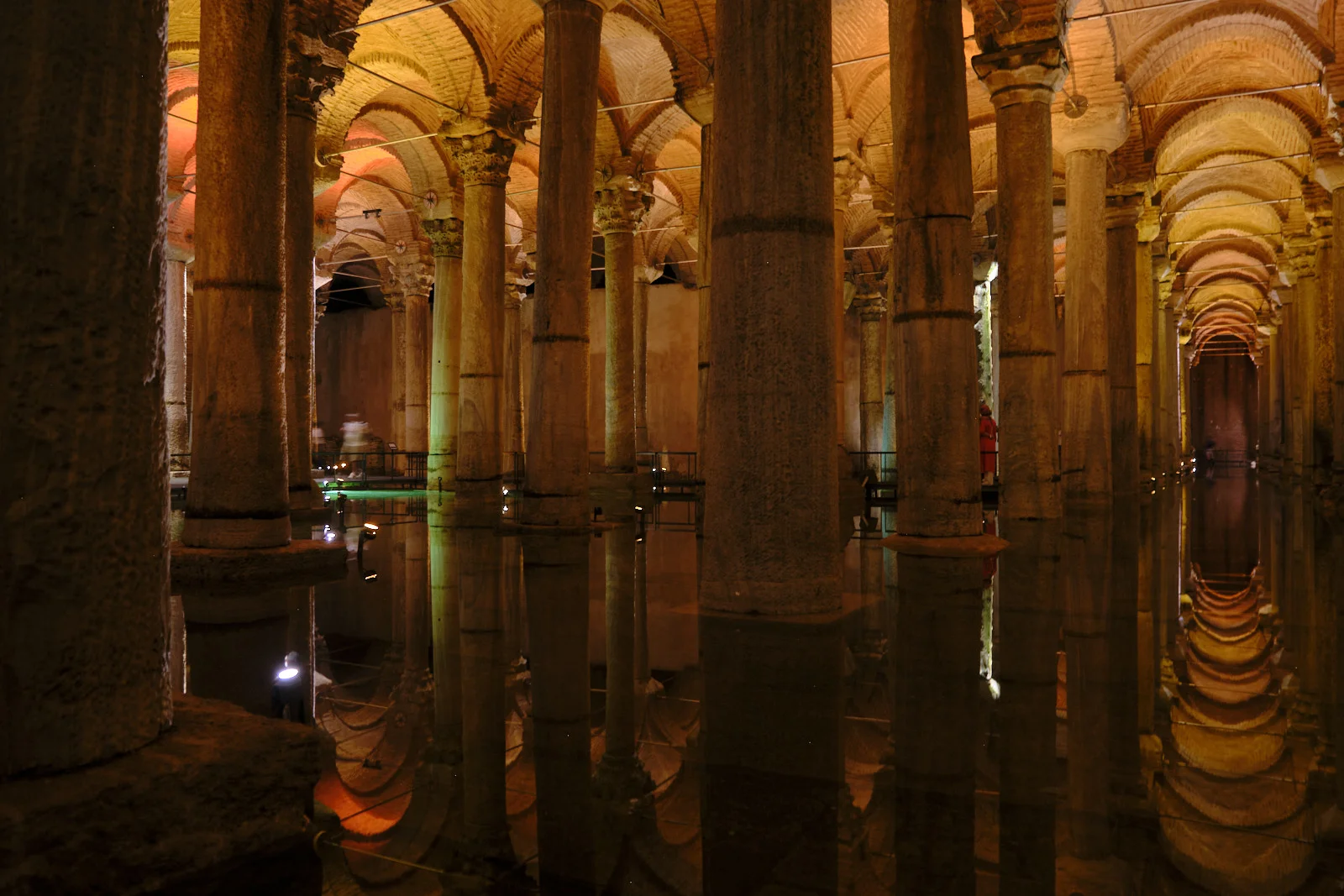 Many stone columns rise from reflective water under brick vaults in the Basilica Cistern, with a person in red on a walkway.