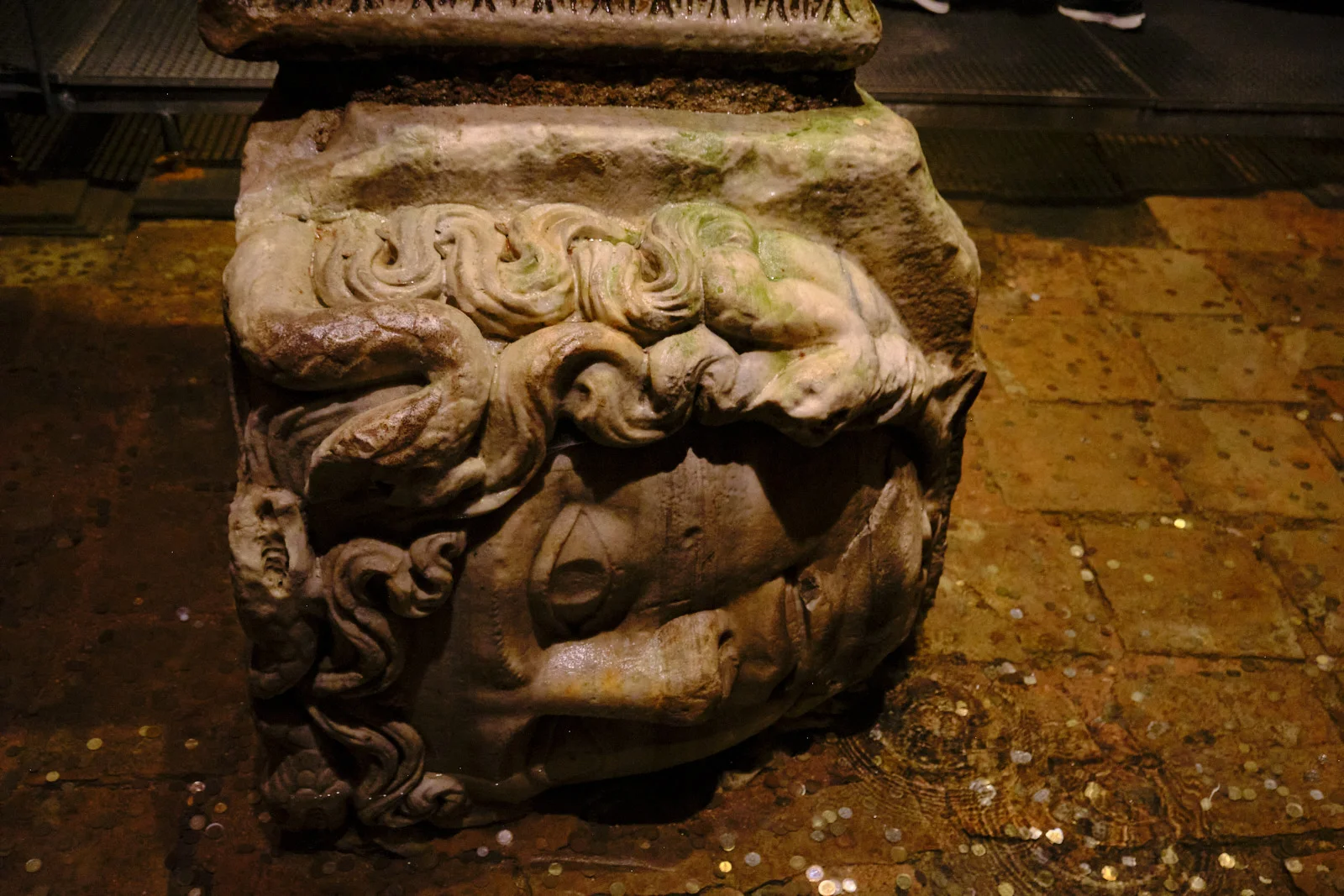 A sideways carved Medusa head with snake hair forms a wet column base, with coins on the floor around it.