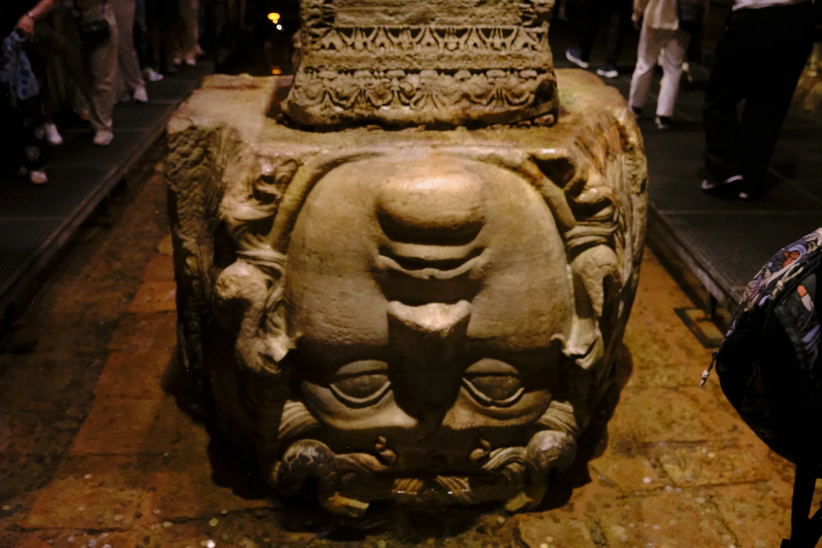 An upside-down carved Medusa head supports a column base while visitors stand on the walkway behind it.