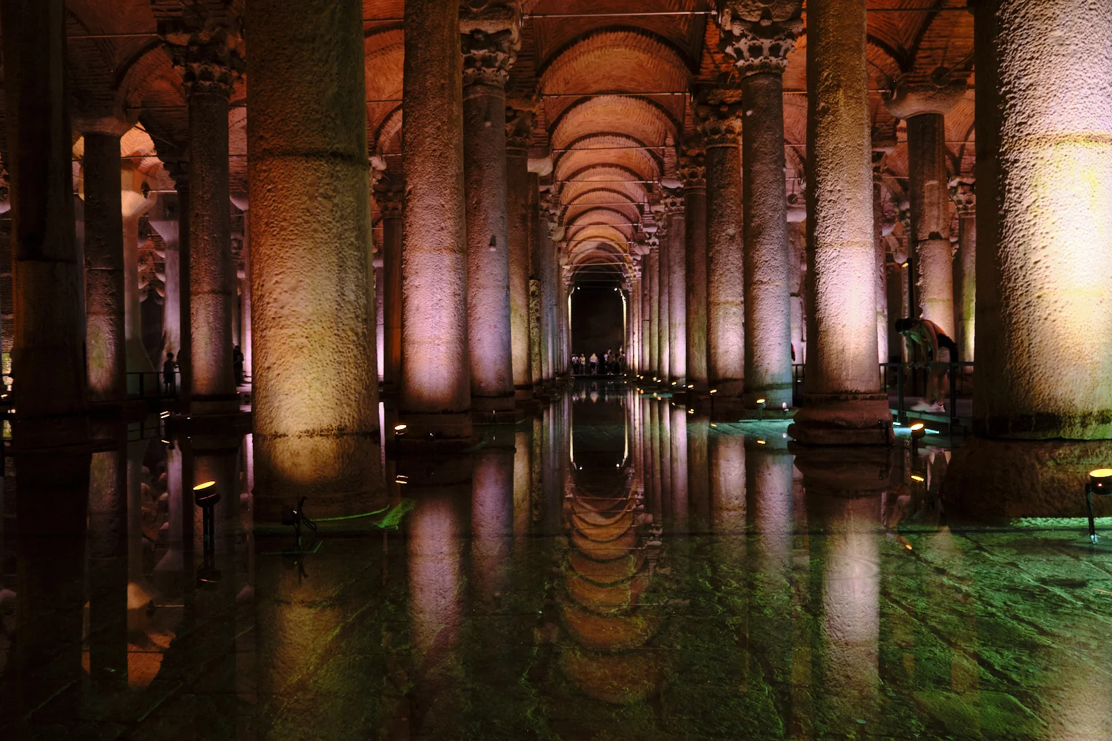 Rows of stone columns and brick arches in the Basilica Cistern, reflected in shallow water under warm and green lights, with a visitor at the right.