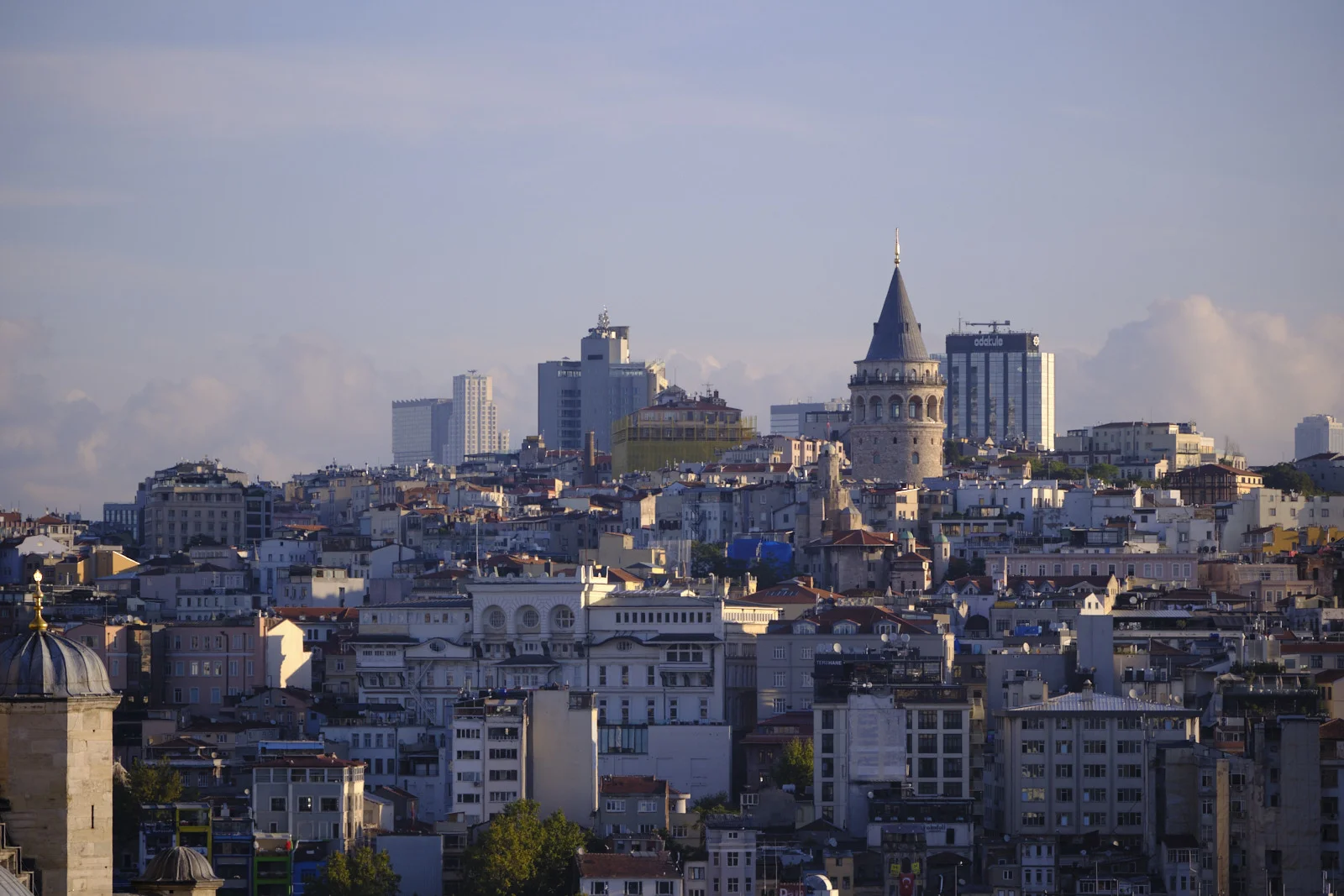 A hazy view across Istanbul rooftops toward the Galata Tower, with modern high-rises behind it and a gilded finial in the lower left foreground.