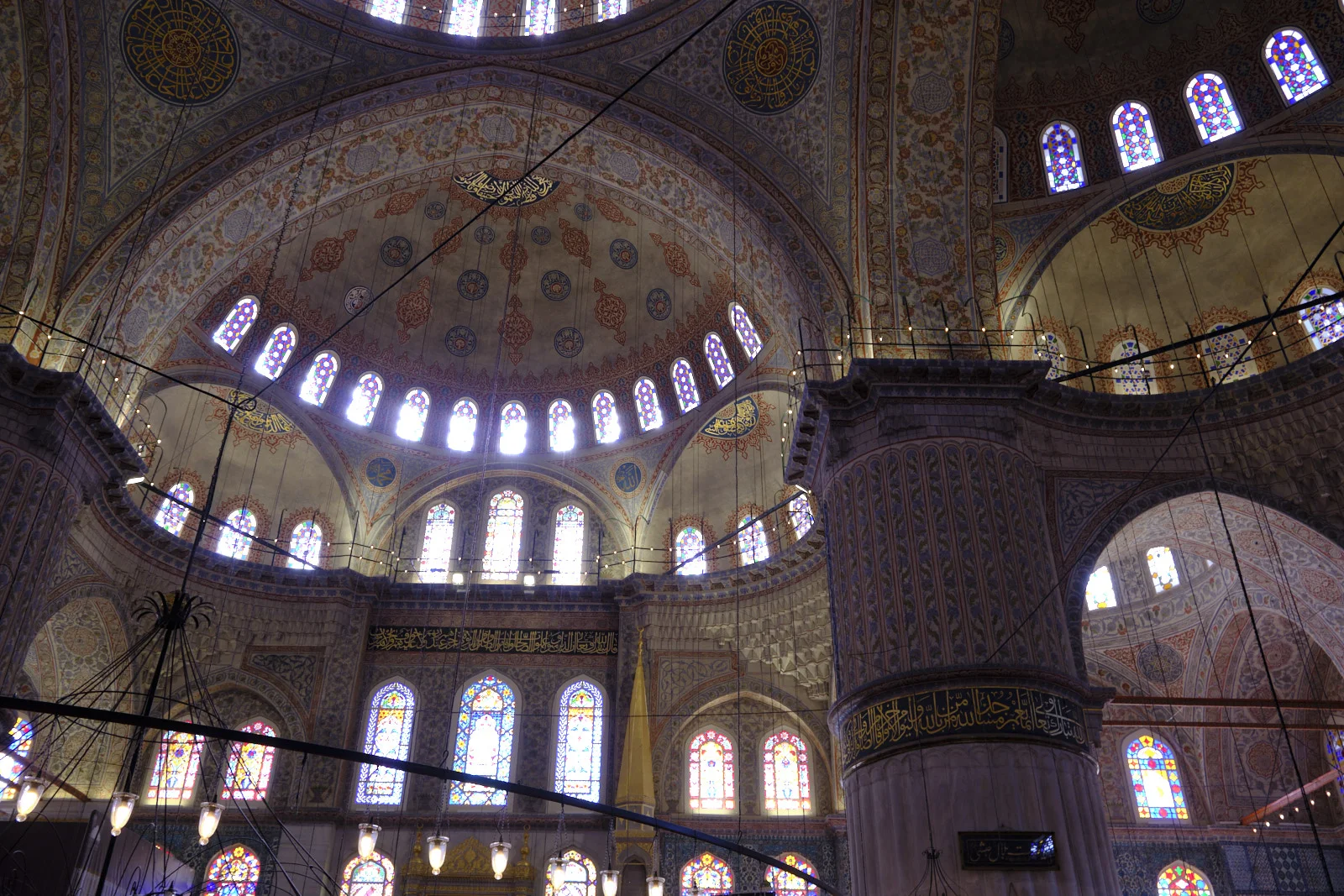 The Blue Mosque interior, with stained-glass windows, painted arches, hanging lamps, and a large stone pier.
